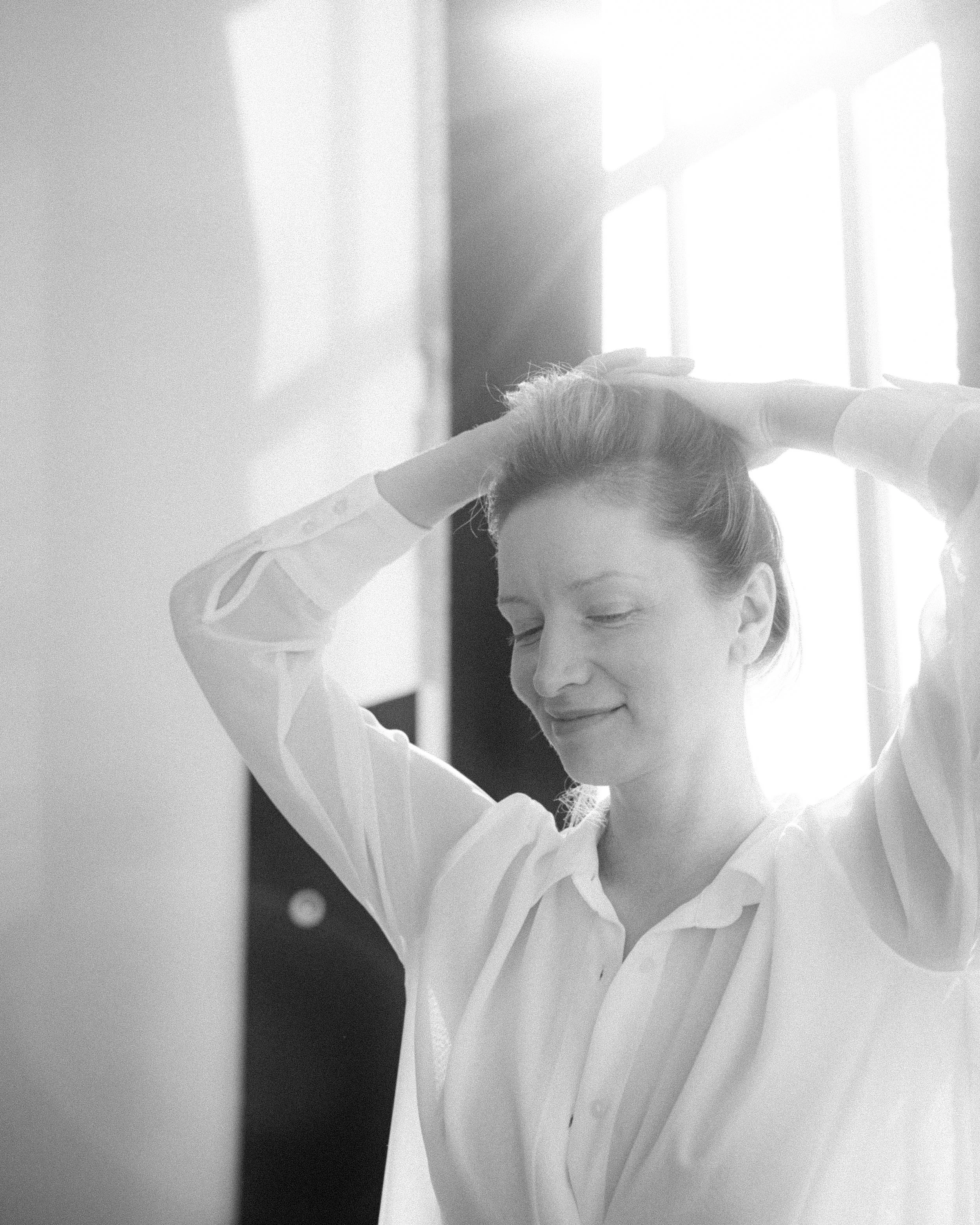 A woman in a white shirt adjusting her hair near a window with sunlight streaming in.