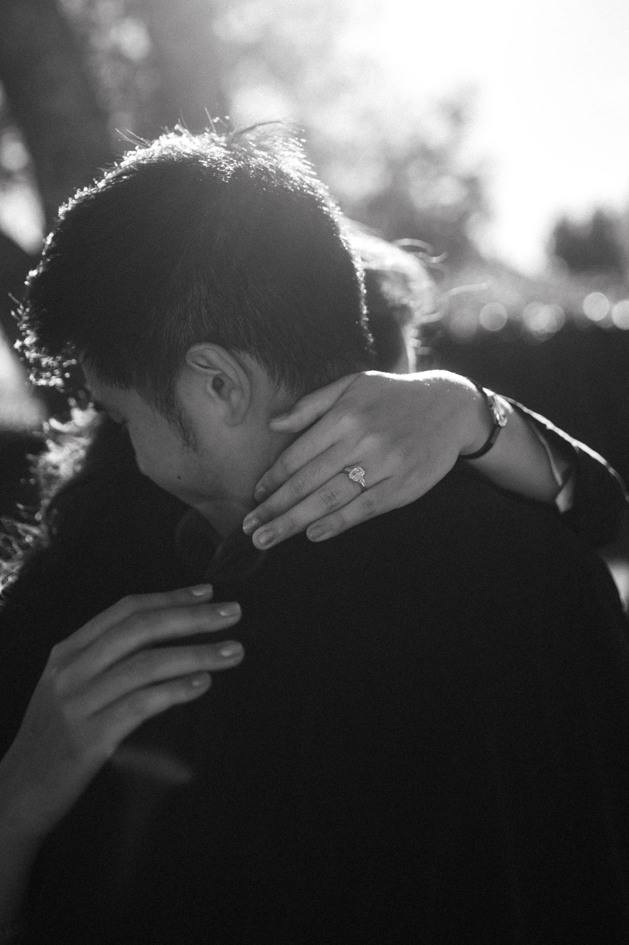 A black and white photo of two people hugging outdoors with sunlight in the background, one person wearing a ring on their finger.