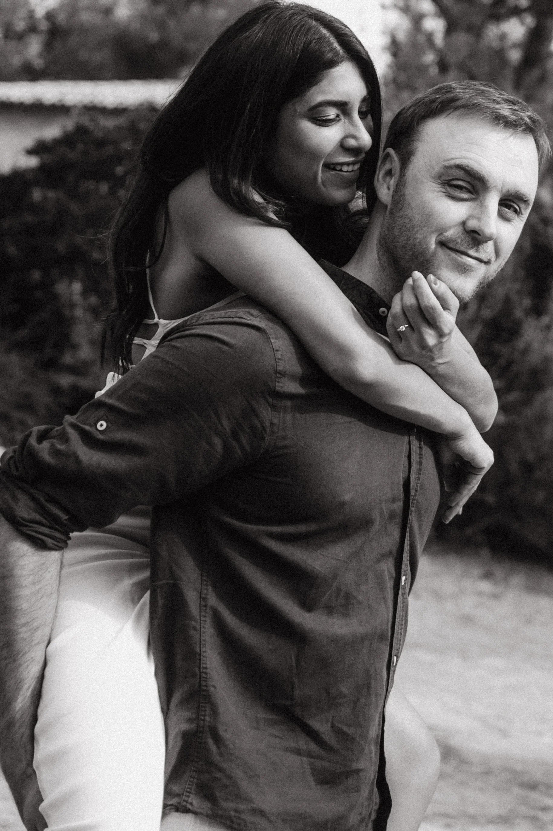 Black and white photo of a man giving a piggyback ride to a woman outdoors, both smiling and making eye contact with the camera.
