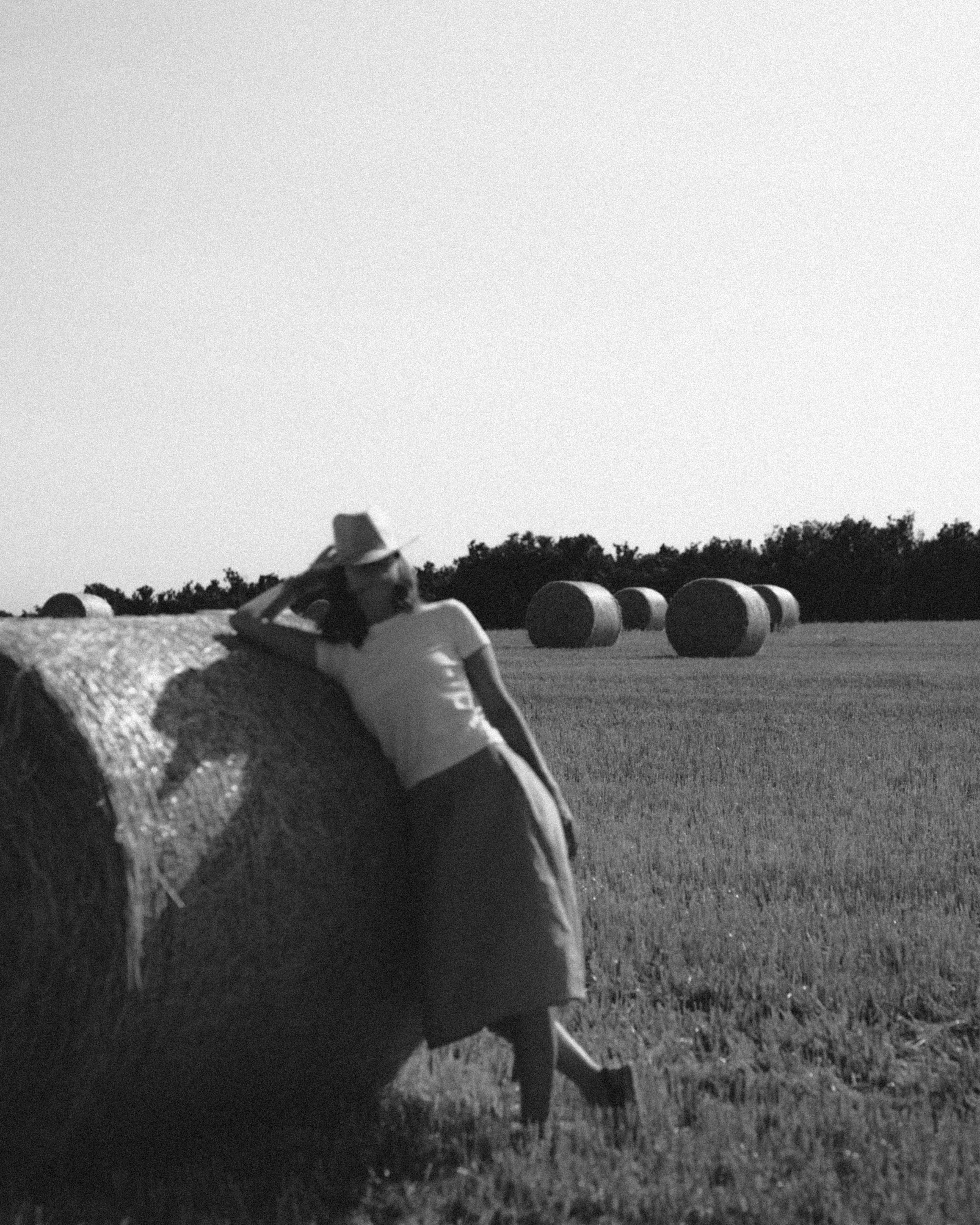 A woman in a hat leaning on a large hay bale in a field with more hay bales in the distance, black and white photo.