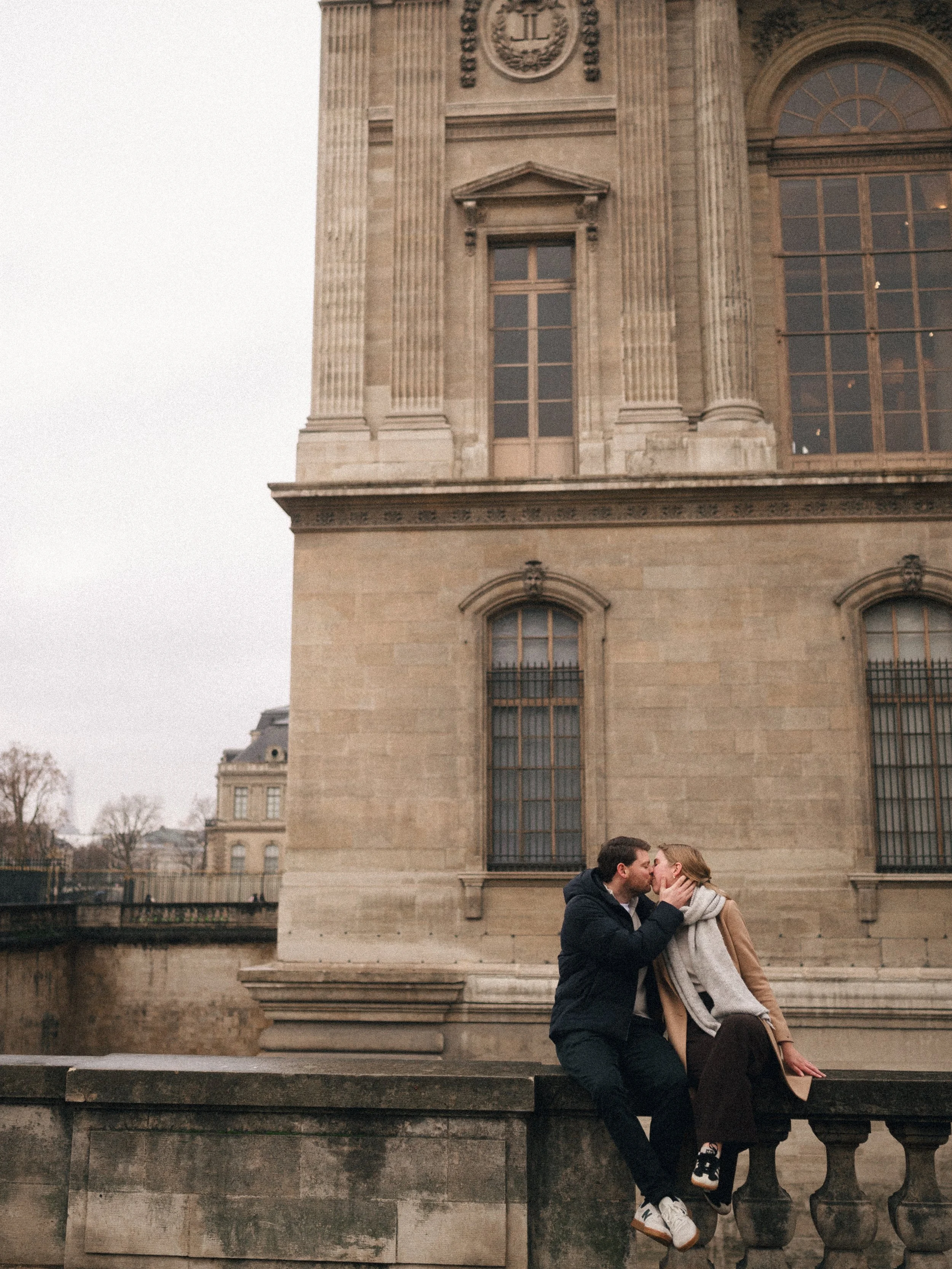 A couple kissing on a stone ledge in front of a historic building with large windows in an urban setting.