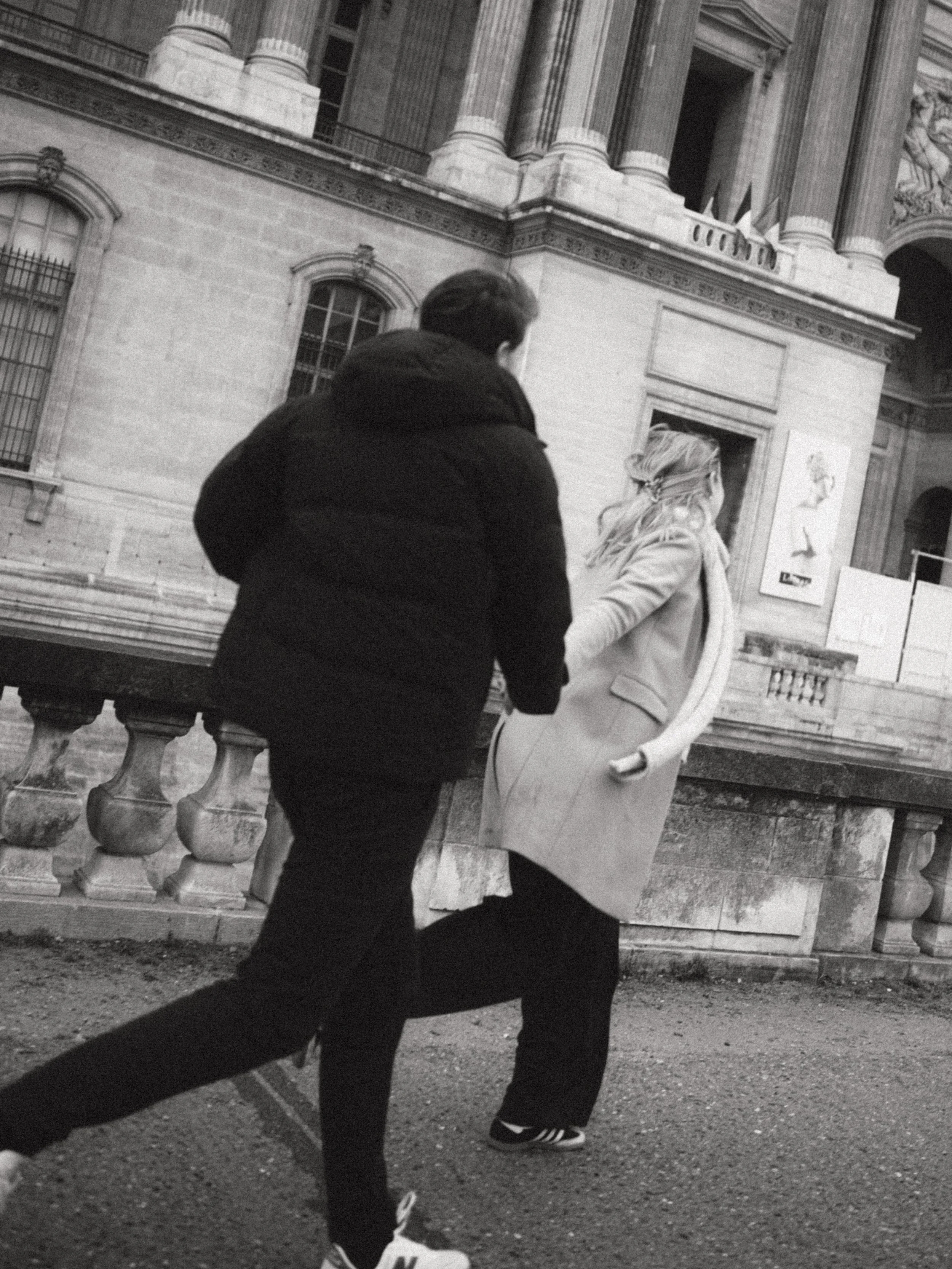 A black-and-white photo of a man and woman walking past a building with classical architecture, including columns and ornate details.