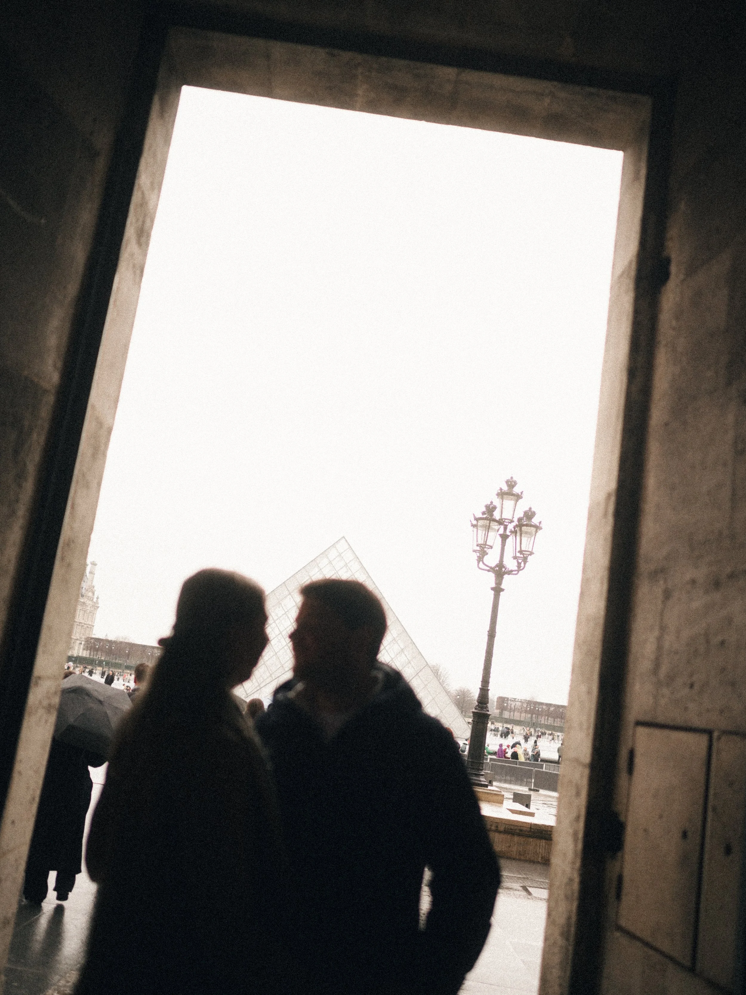 Silhouettes of a man and a woman standing close together, seen through an arched doorway, with the Louvre Pyramid and a streetlamp in the background, in Paris.