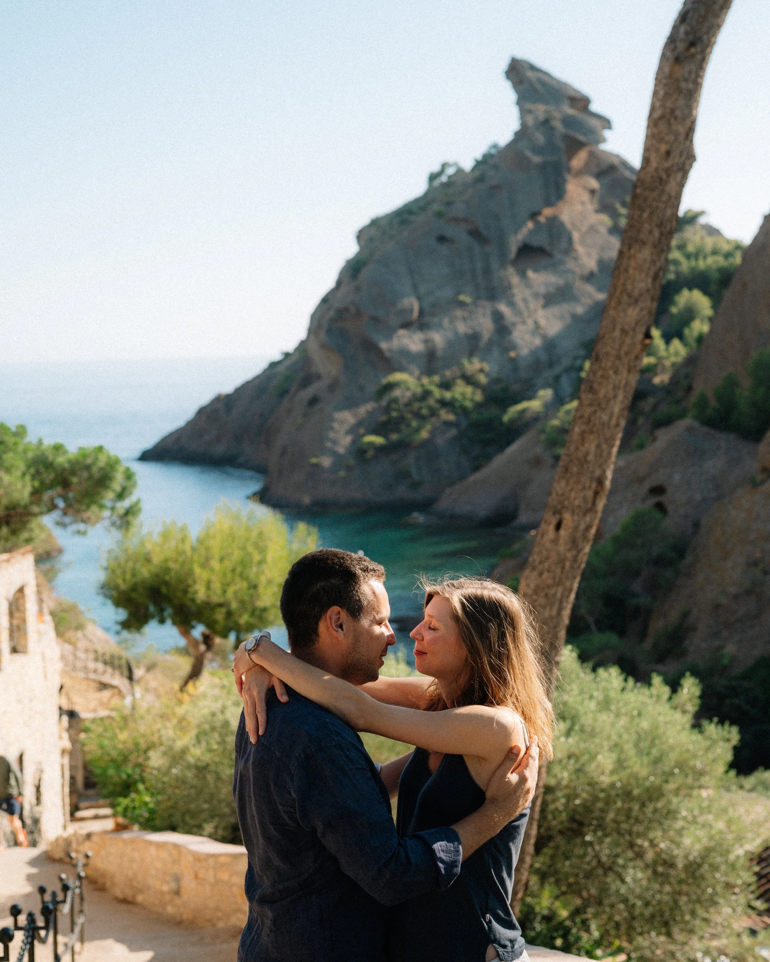 A couple embraces by a scenic coastal landscape with rocky hills, trees, and a body of water in the background.