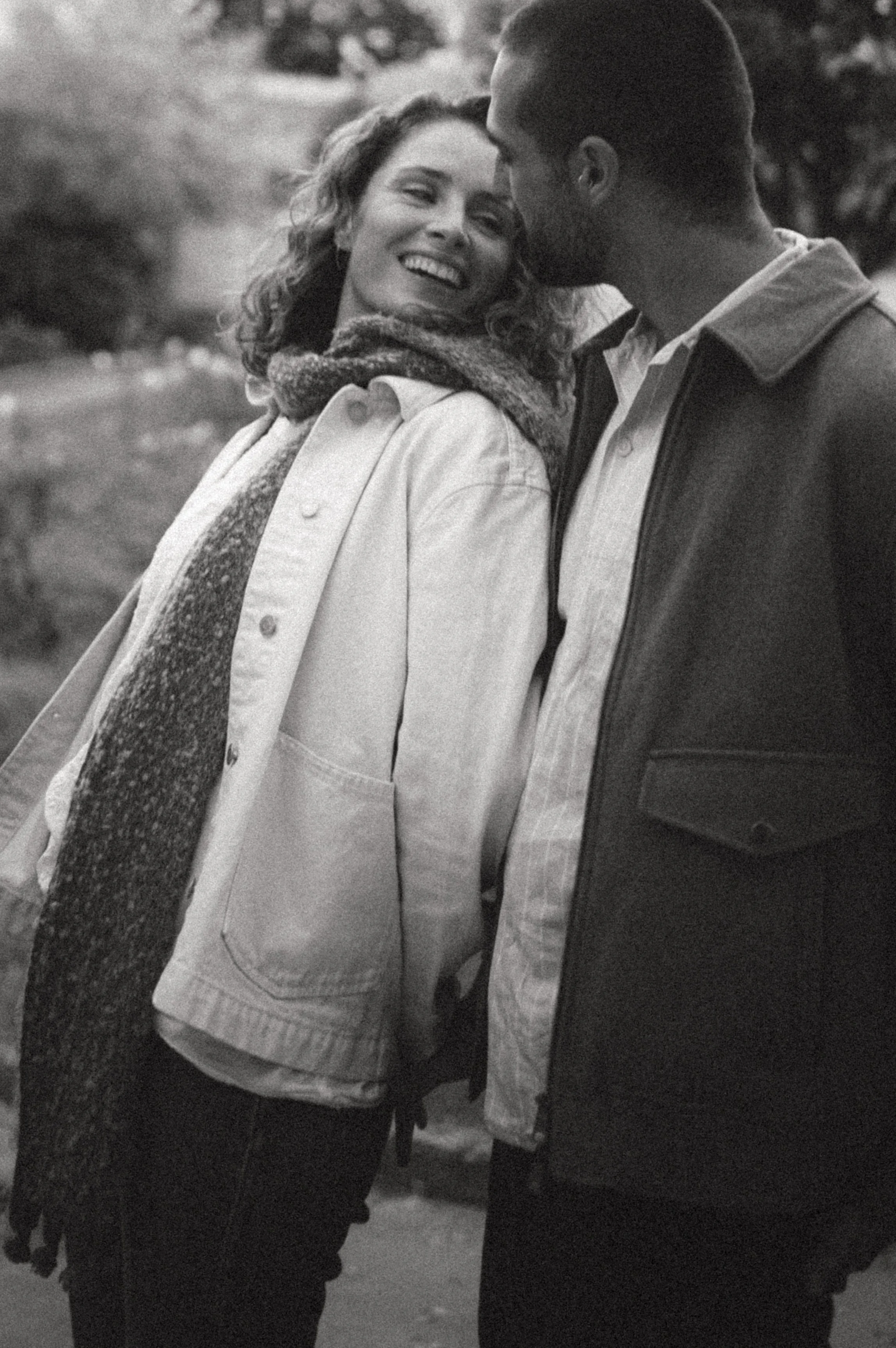 A black and white photo of a smiling woman and a bearded man standing close outdoors. The woman is wearing a light jacket and a scarf, and the man is wearing a dark jacket over a light shirt.