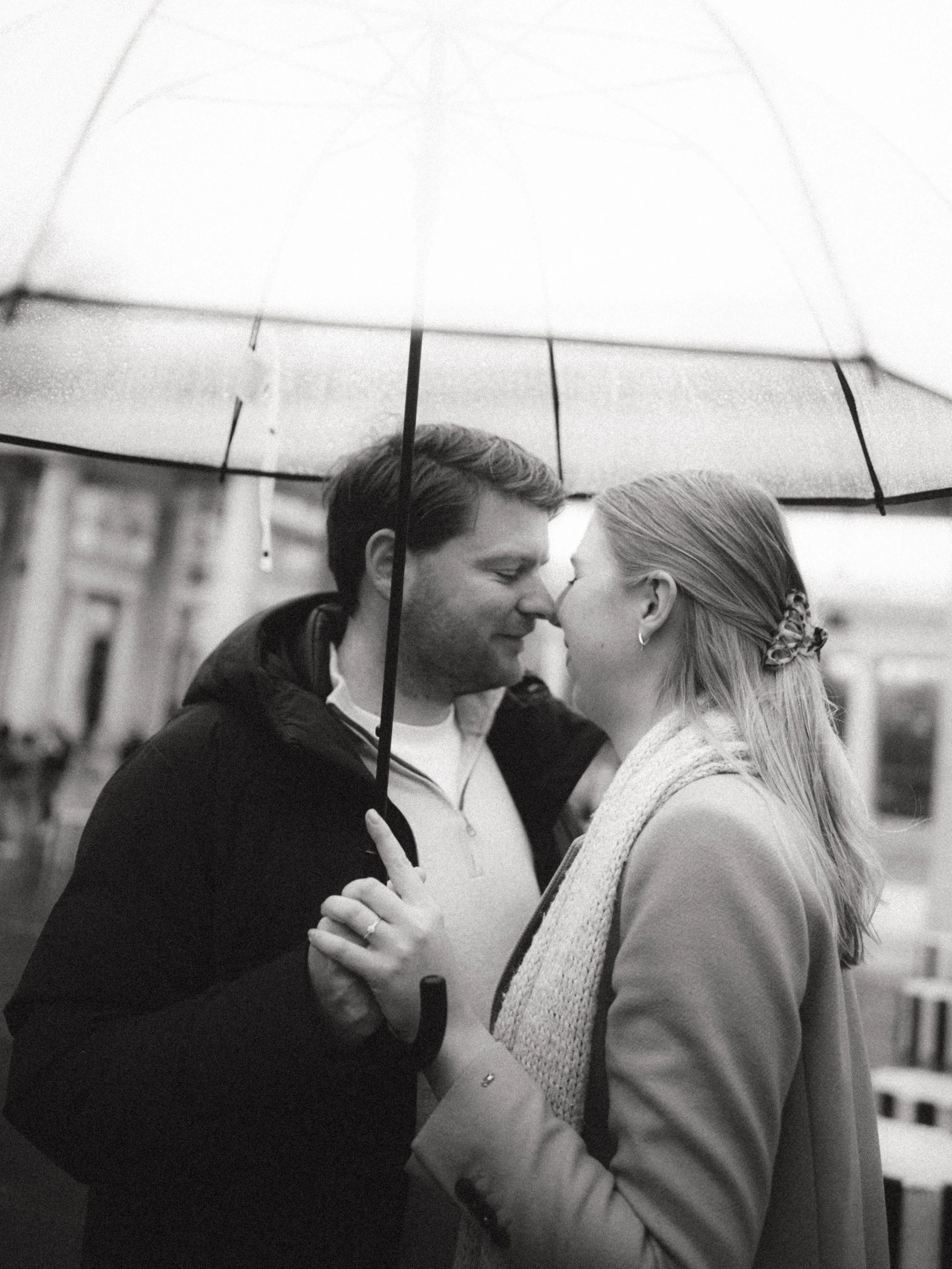 A black and white photo of a couple sharing a close moment under an umbrella on a rainy day, facing each other with noses touching.