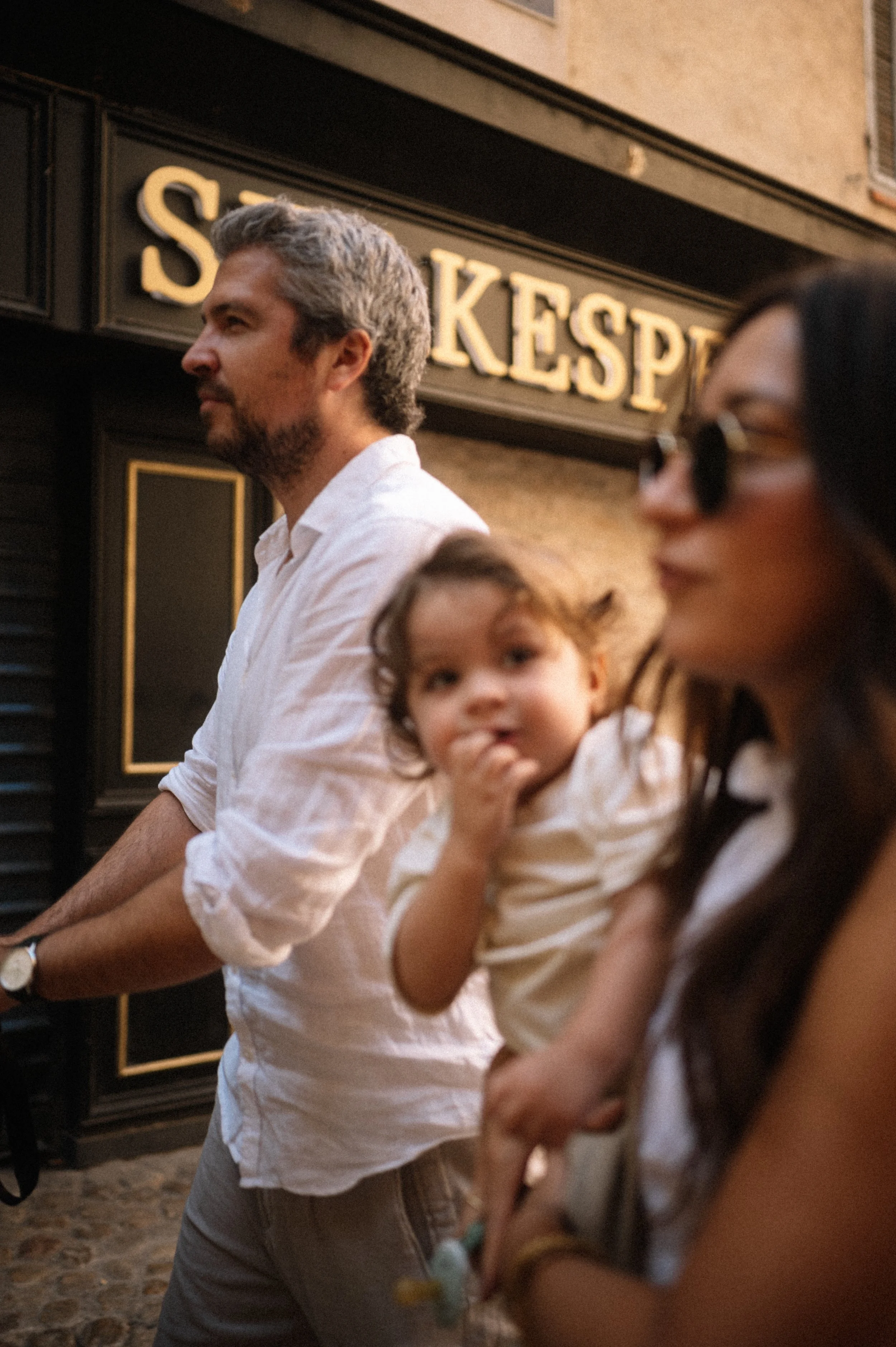 A man with gray hair and a beard wearing a white shirt, a woman with dark hair and sunglasses, and a little girl with curly hair and a striped shirt standing outdoors near a building with a sign that reads 'SKEP' in gold letters.