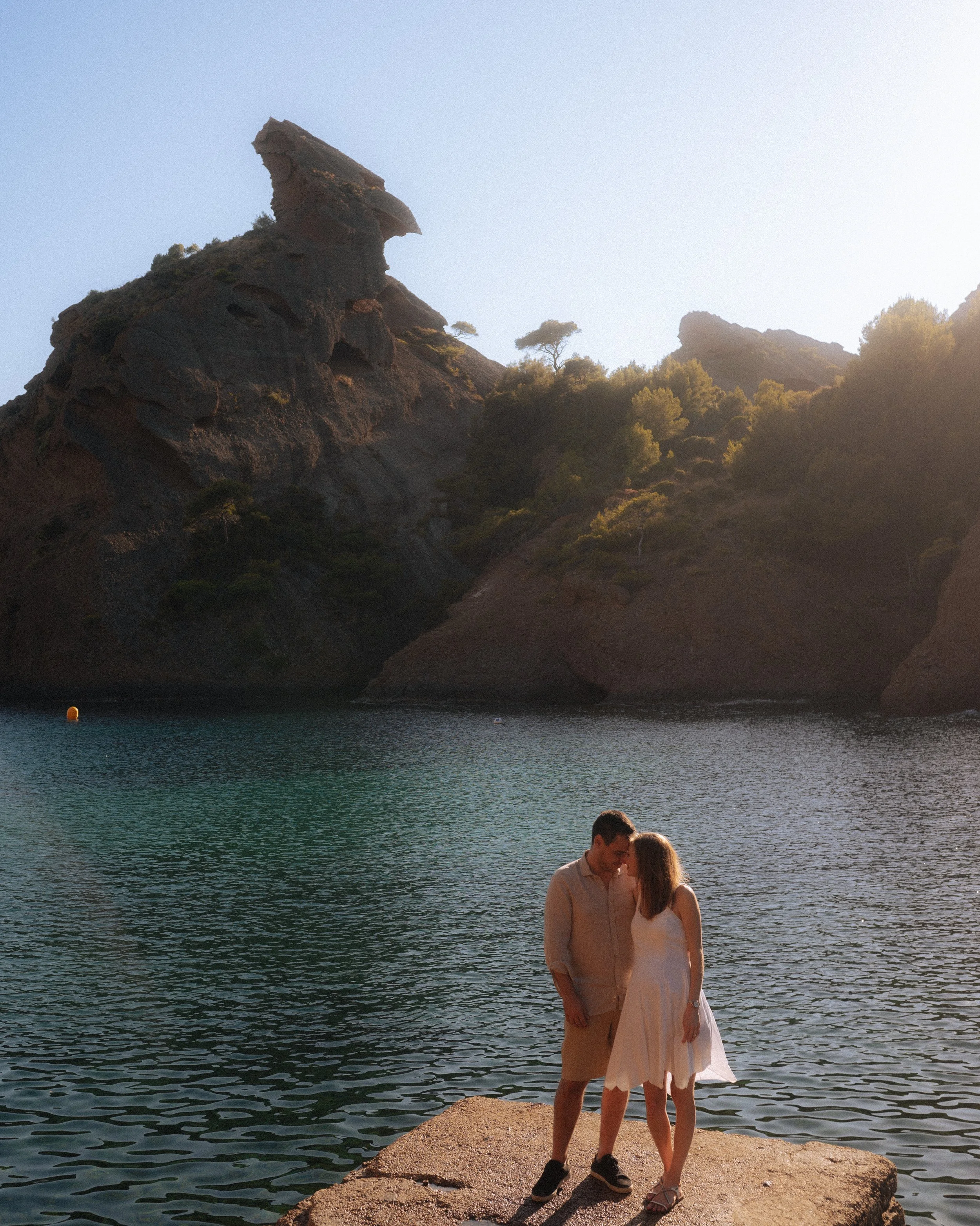A couple standing on a rocky ledge by a lake with a large rock formation and trees in the background during sunset or sunrise.