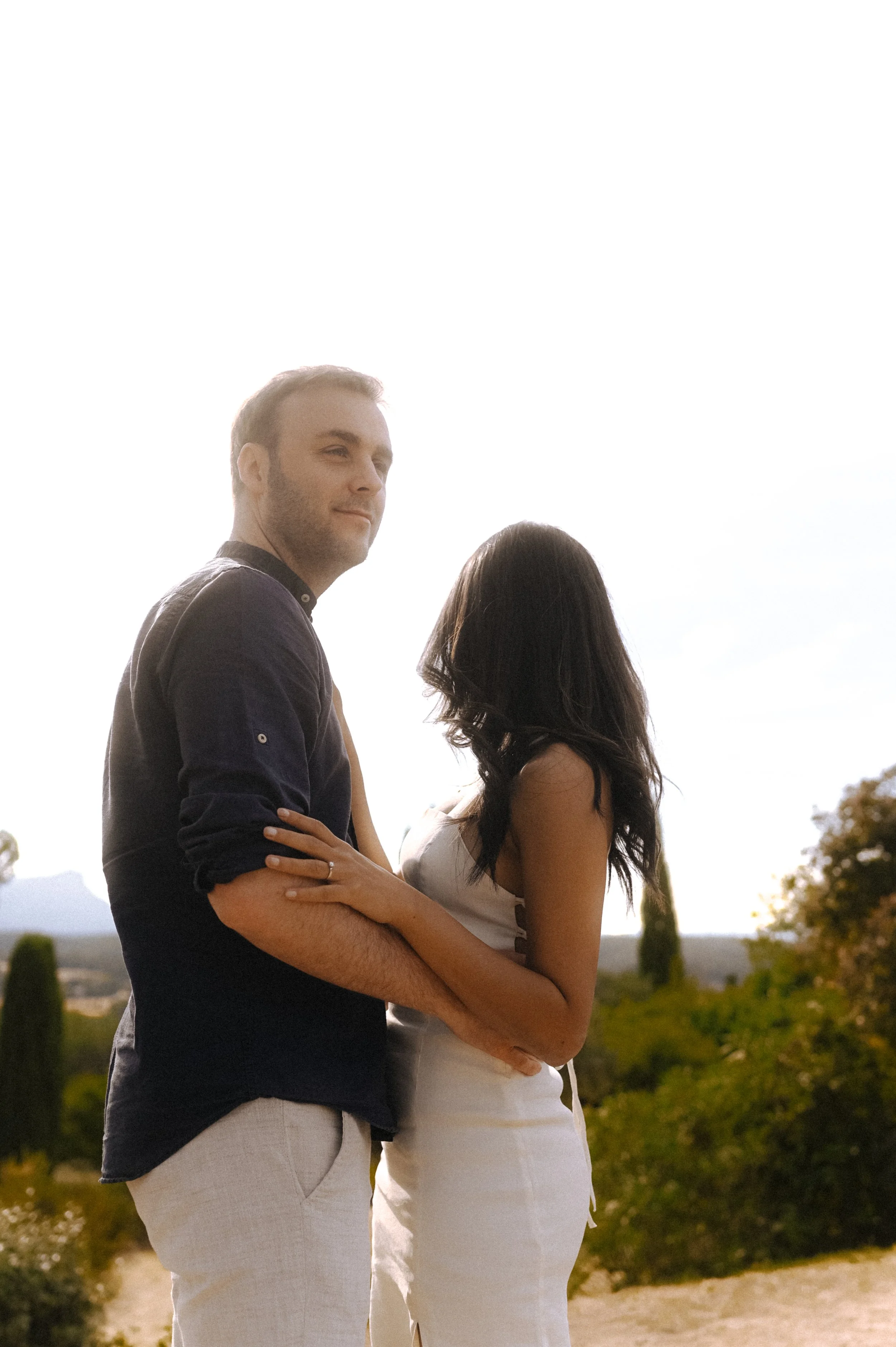 A man and woman standing outdoors with a scenic landscape behind them, facing away from each other but close together.