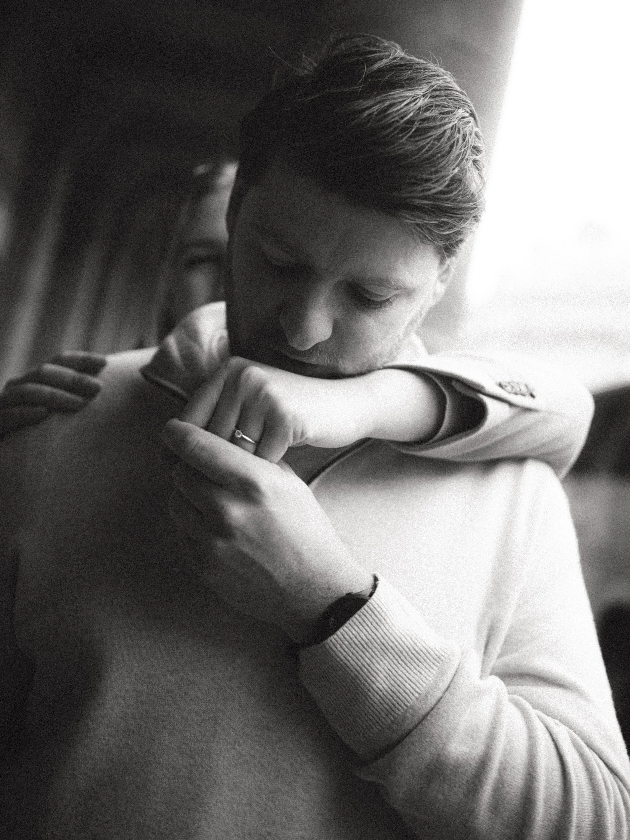 A black and white photograph of a man with closed eyes, gently holding and resting his head on a woman's arm. The woman is wearing a light-colored sweater and a ring on her finger.