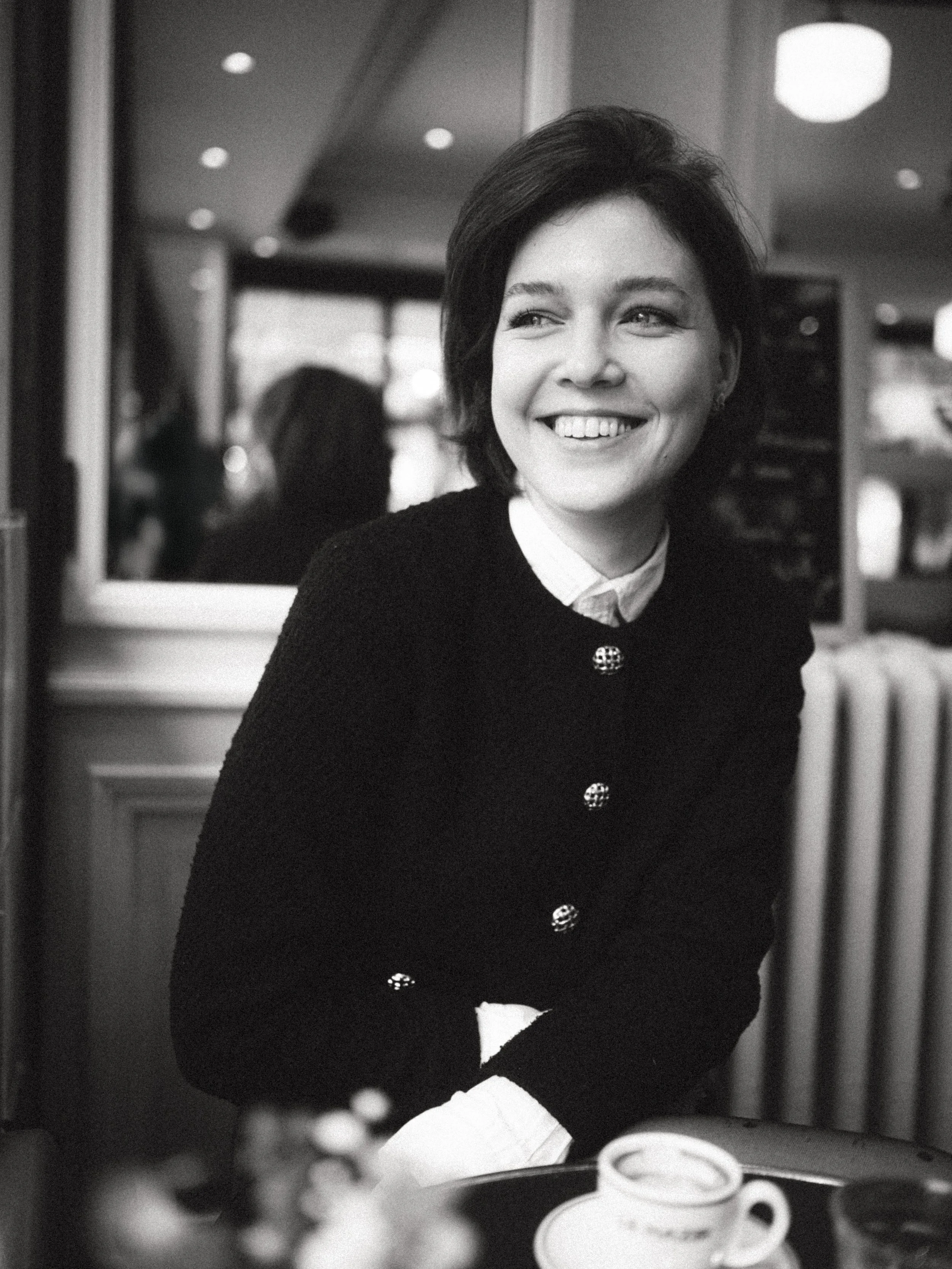 Black and white photo of a woman smiling, seated in a cafe or restaurant, with a cup of coffee on the table in front of her.