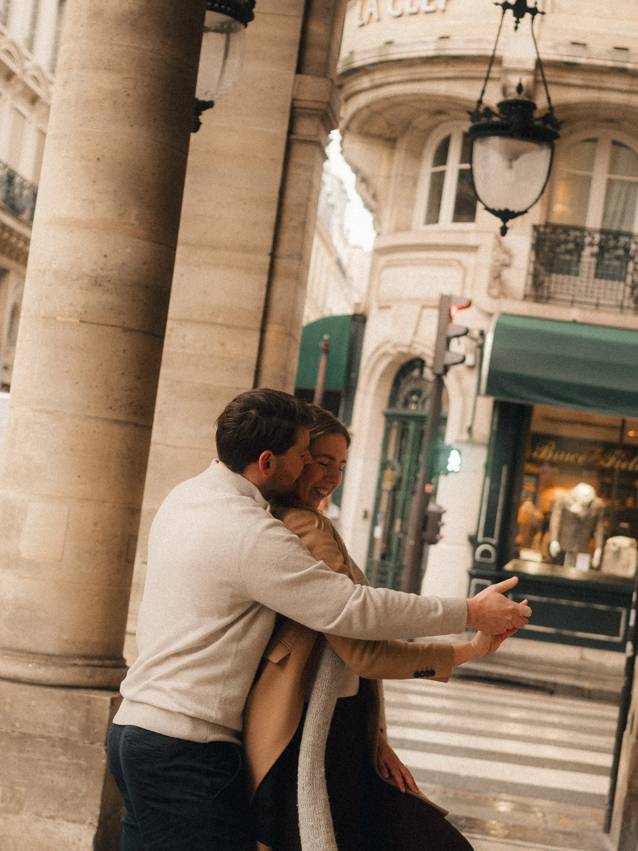A couple is taking a selfie together on a city street, smiling and hugging. The background features historic buildings and streetlamps.