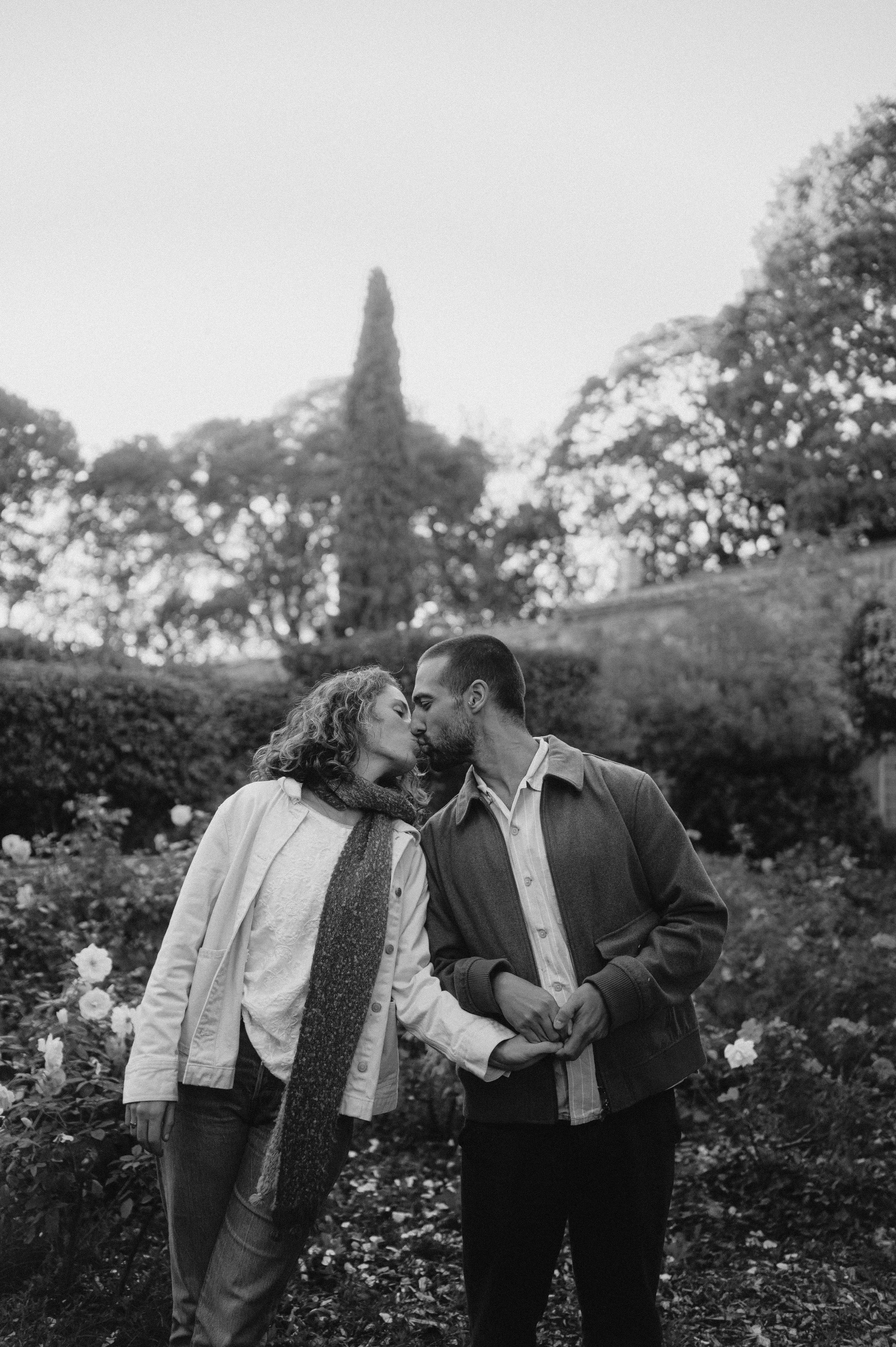 A black and white photo of a couple sharing a kiss in a park with flowers and trees in the background.