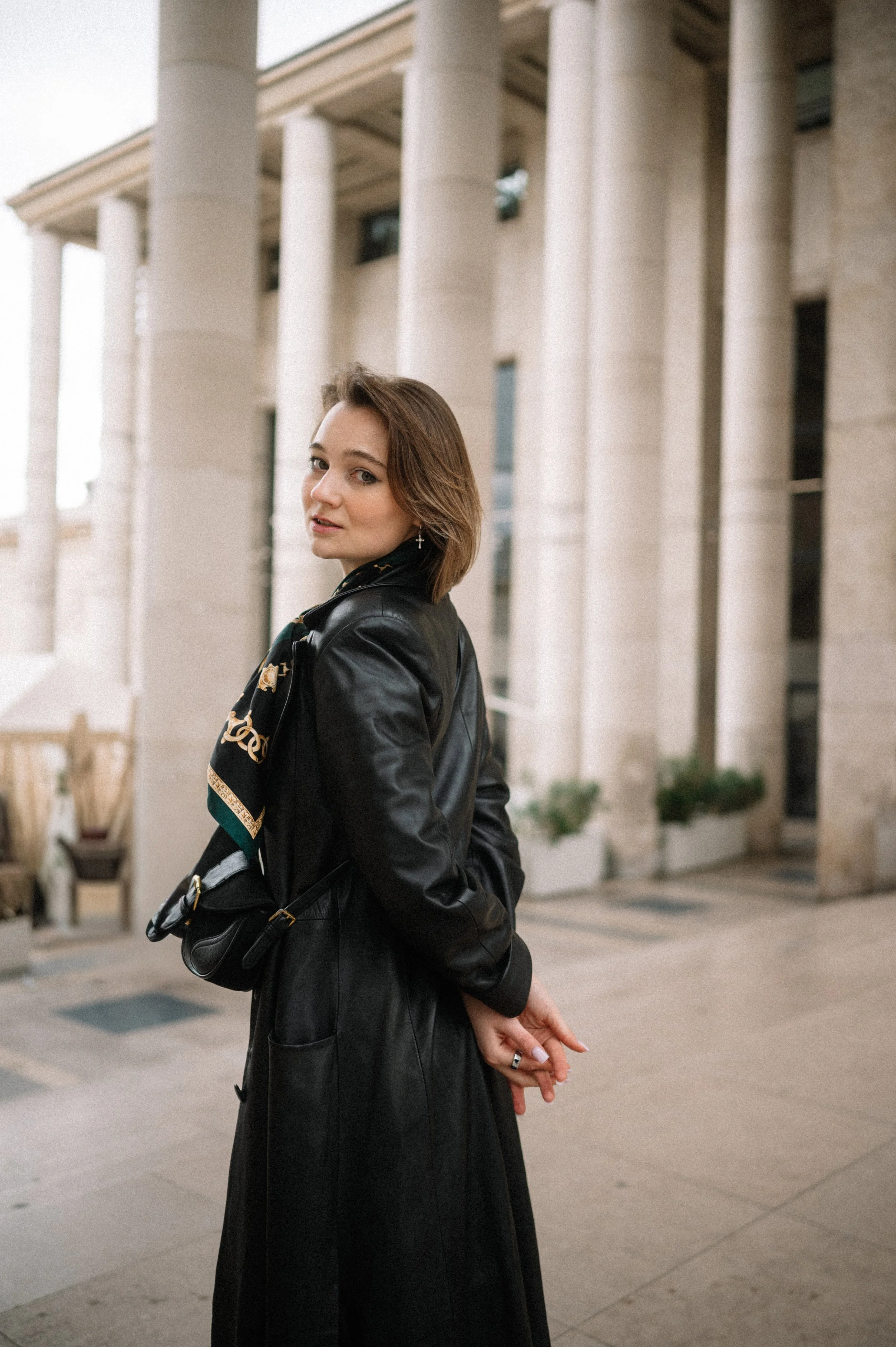A woman in a black leather trench coat standing outdoors in front of a building with large columns, looking at the camera.