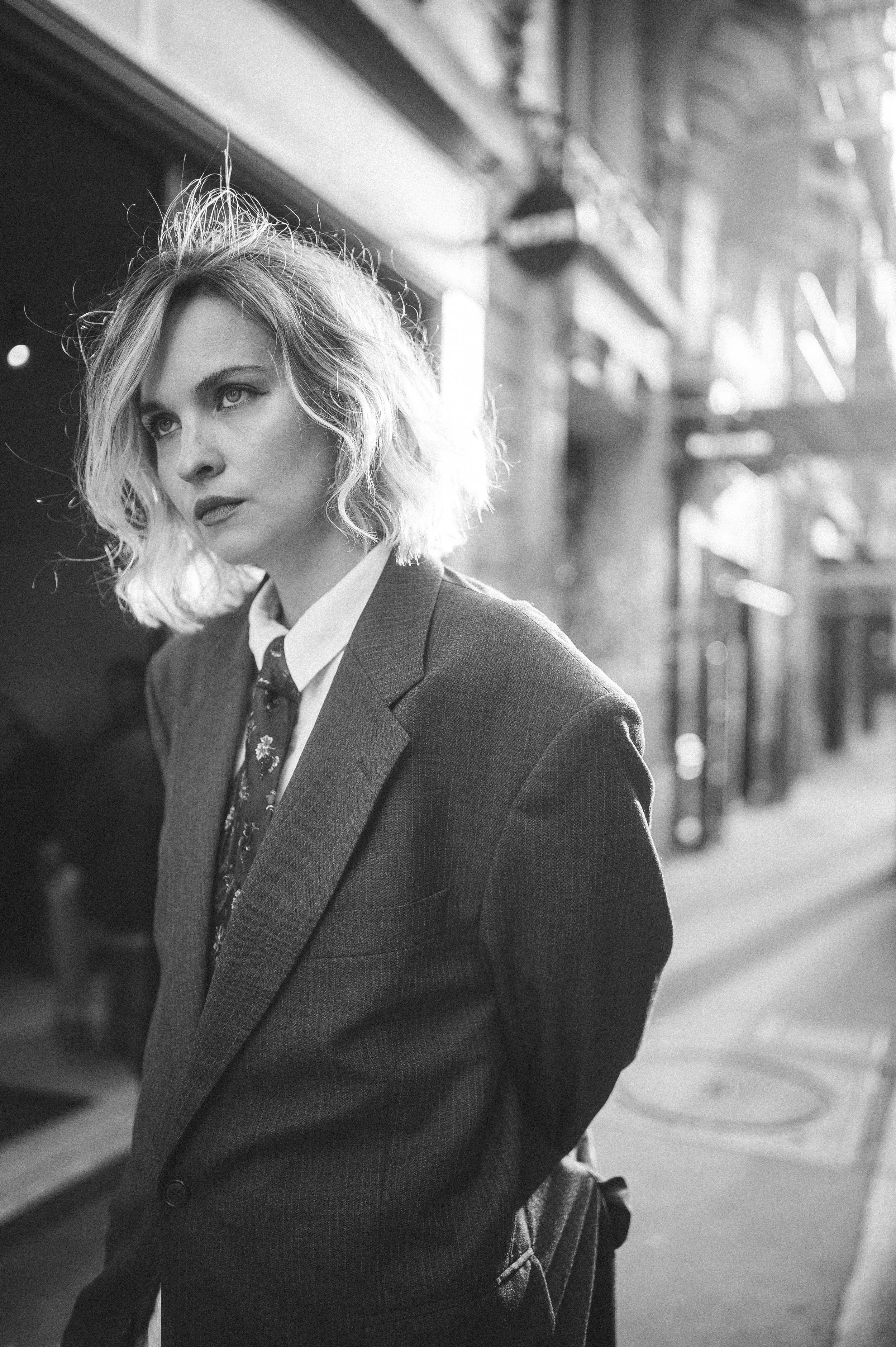 A black and white photo of a woman with wavy blond hair, wearing a suit jacket, shirt, and tie, standing on an urban sidewalk with blurred storefronts and streetlights in the background.