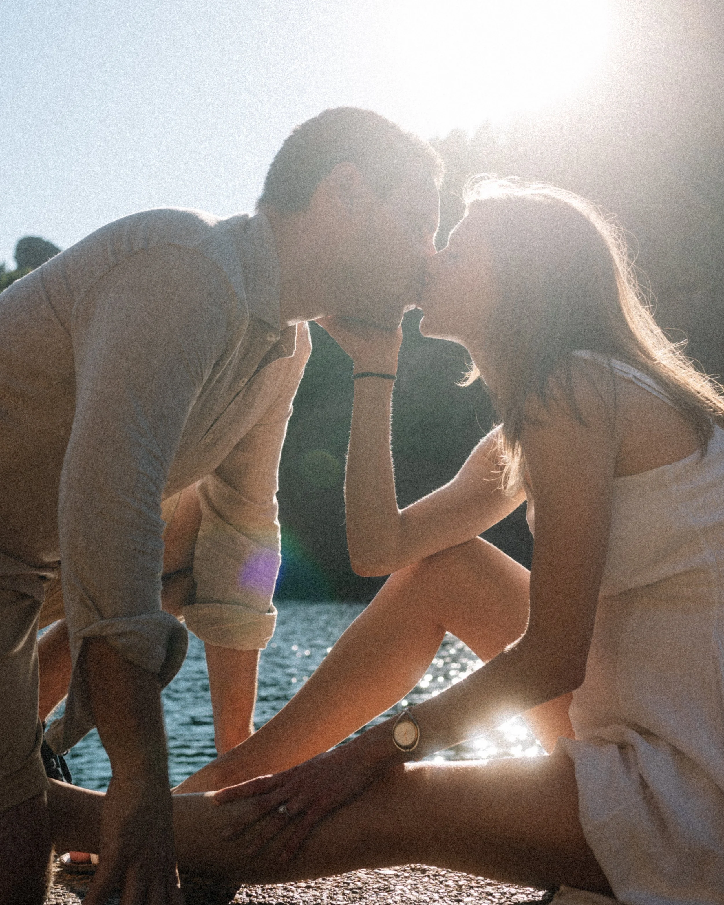 A couple kissing outdoors near a body of water, sunlight shining behind them, creating a backlit scene.