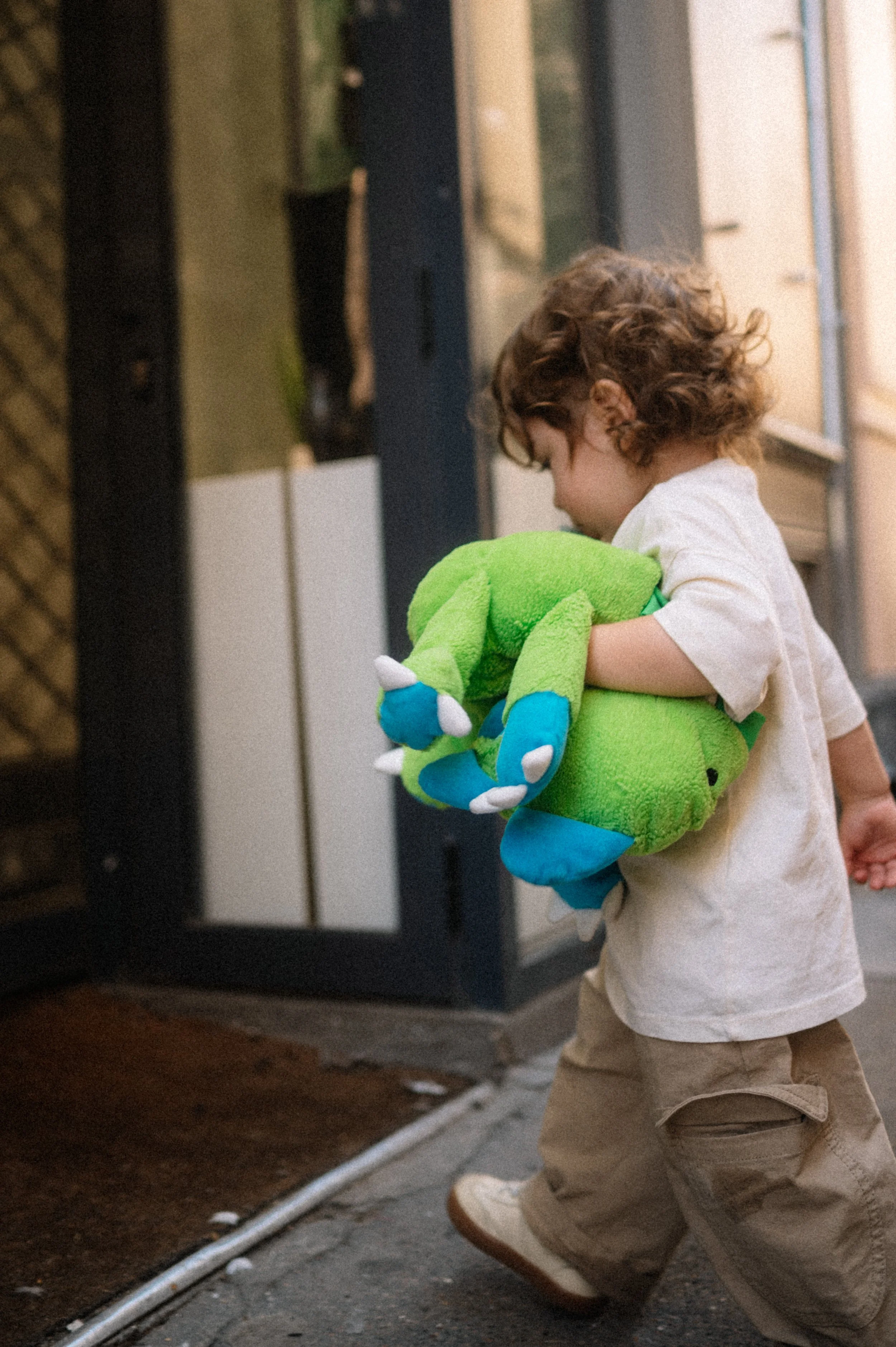 A young child with curly hair wearing a white shirt and beige pants, holding a bright green plush dinosaur toy with blue and white accents.