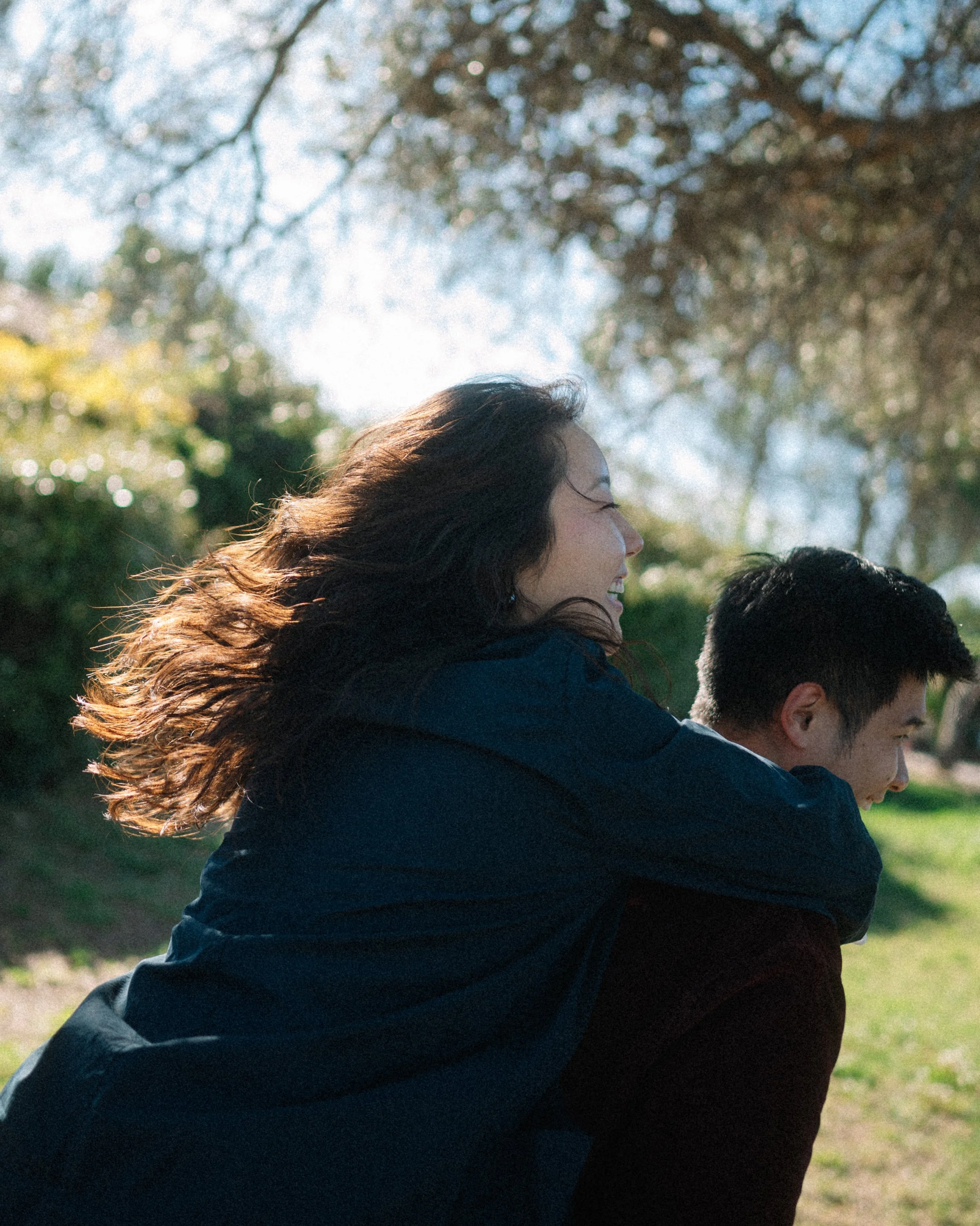 A woman with long, wavy hair smiling and laughing while riding on the back of a man outdoors in a park with trees and sunlight.