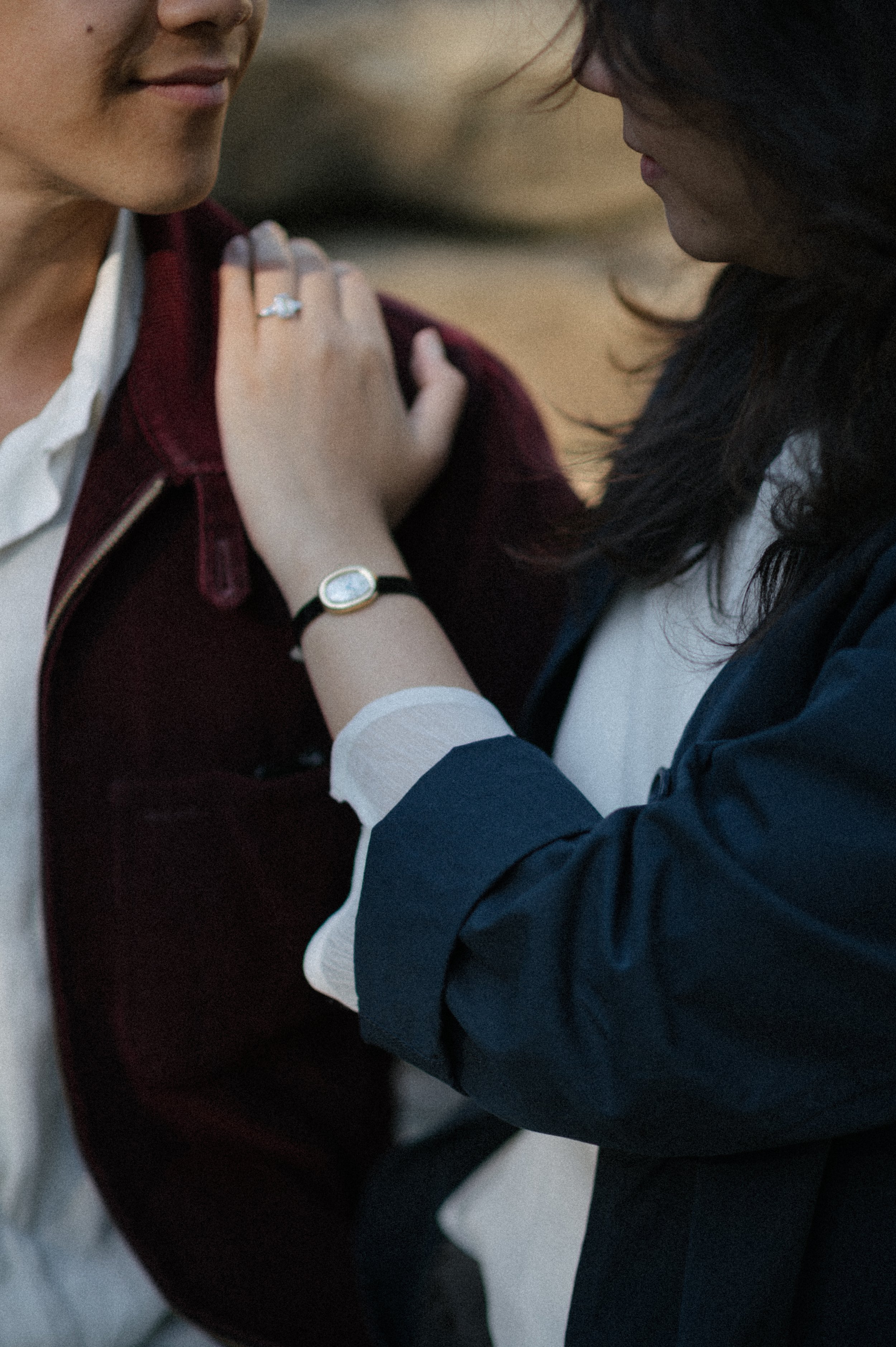 Two women face each other closely, one touching the other's shoulder. One woman wears a ring and a watch, and the background is blurred outdoors.