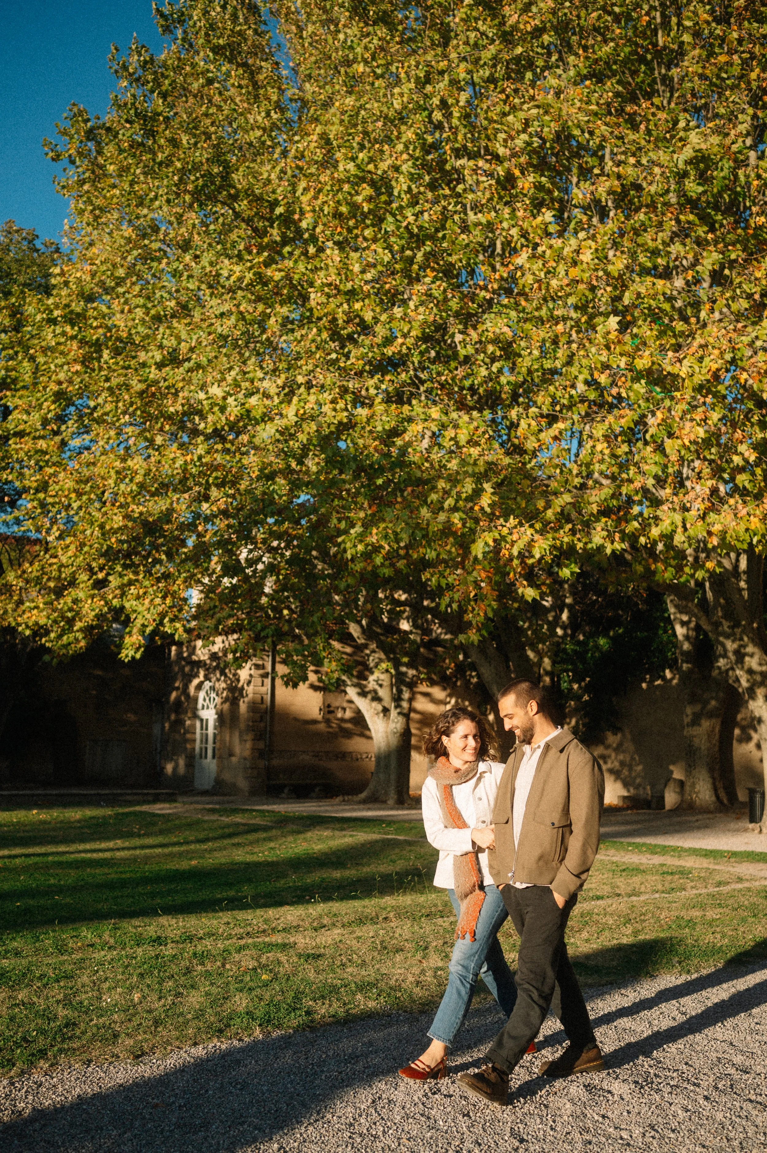 A young couple walking on a gravel path in a park during autumn, holding hands and smiling, with large trees with yellow and green leaves in the background on a sunny day.