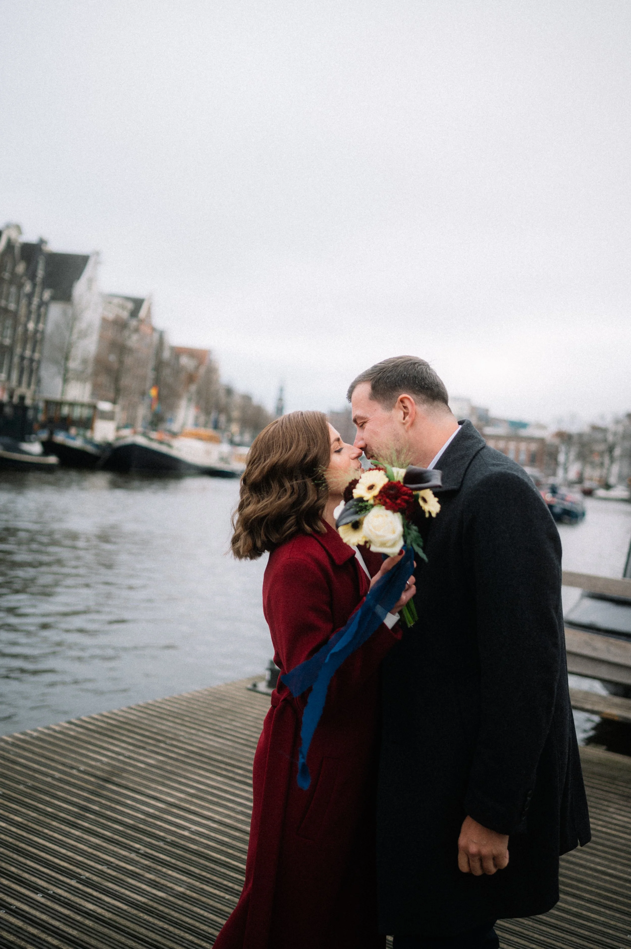 A couple is close together on a wooden dock by a canal, with the woman holding a bouquet of flowers. They are about to kiss in an outdoor city setting, overcast weather.