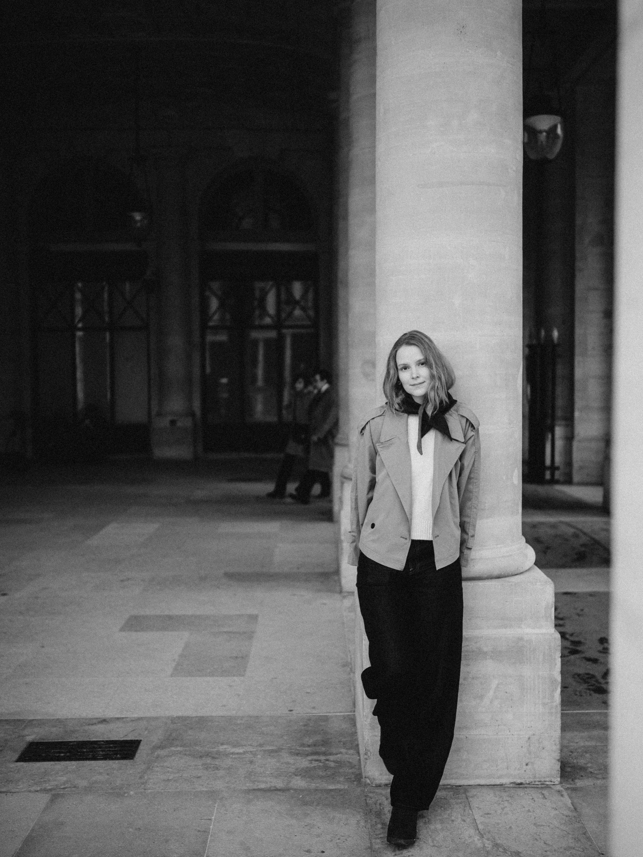 A woman stands leaning against a large column in a city street at night, with a building and a person sitting on a bench in the background, in black and white.