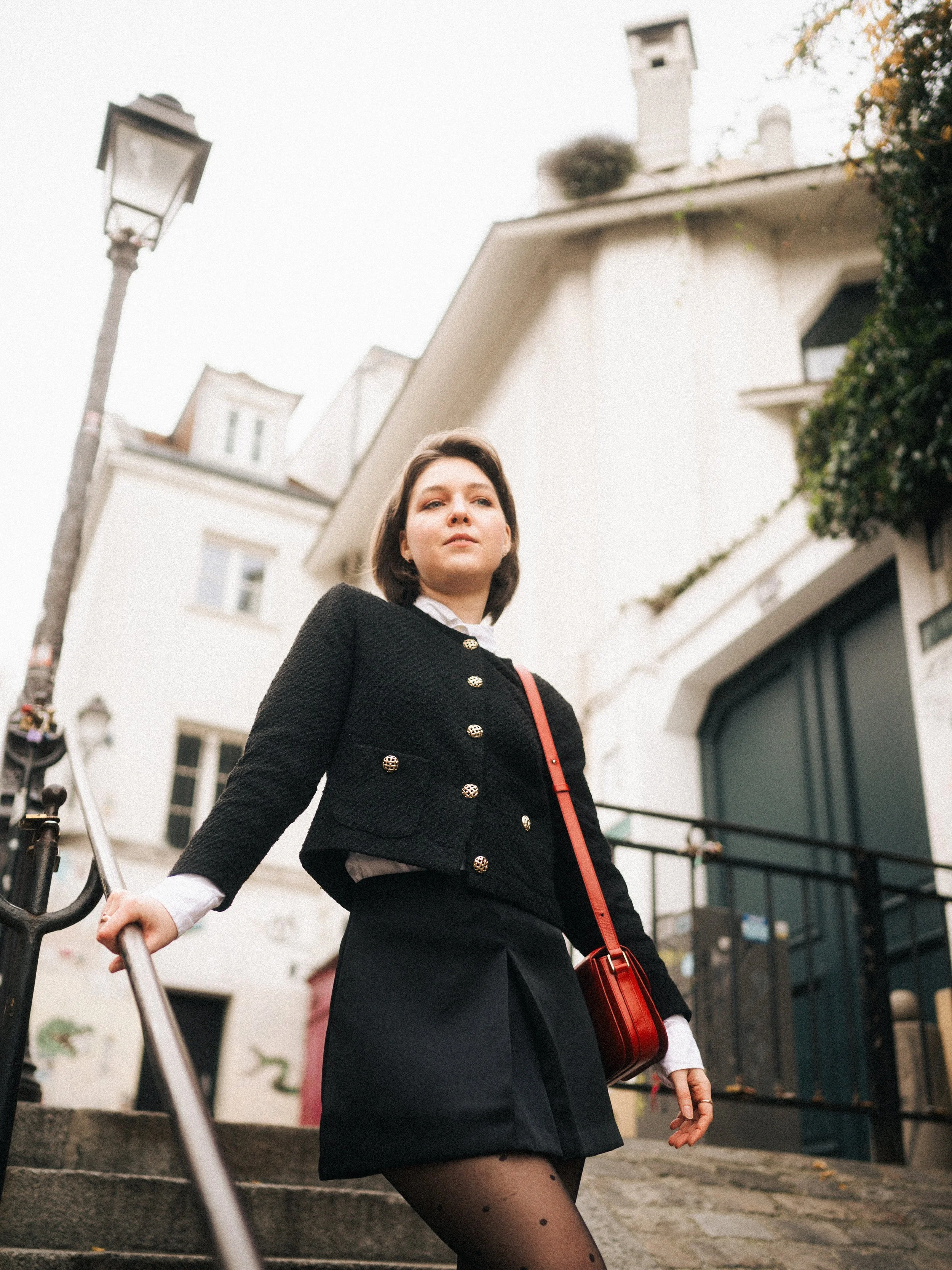Young woman with short brown hair standing on stairs in an urban setting, wearing a black textured jacket with shiny buttons, a black skirt, and polka dot stockings, carrying a red crossbody bag.
