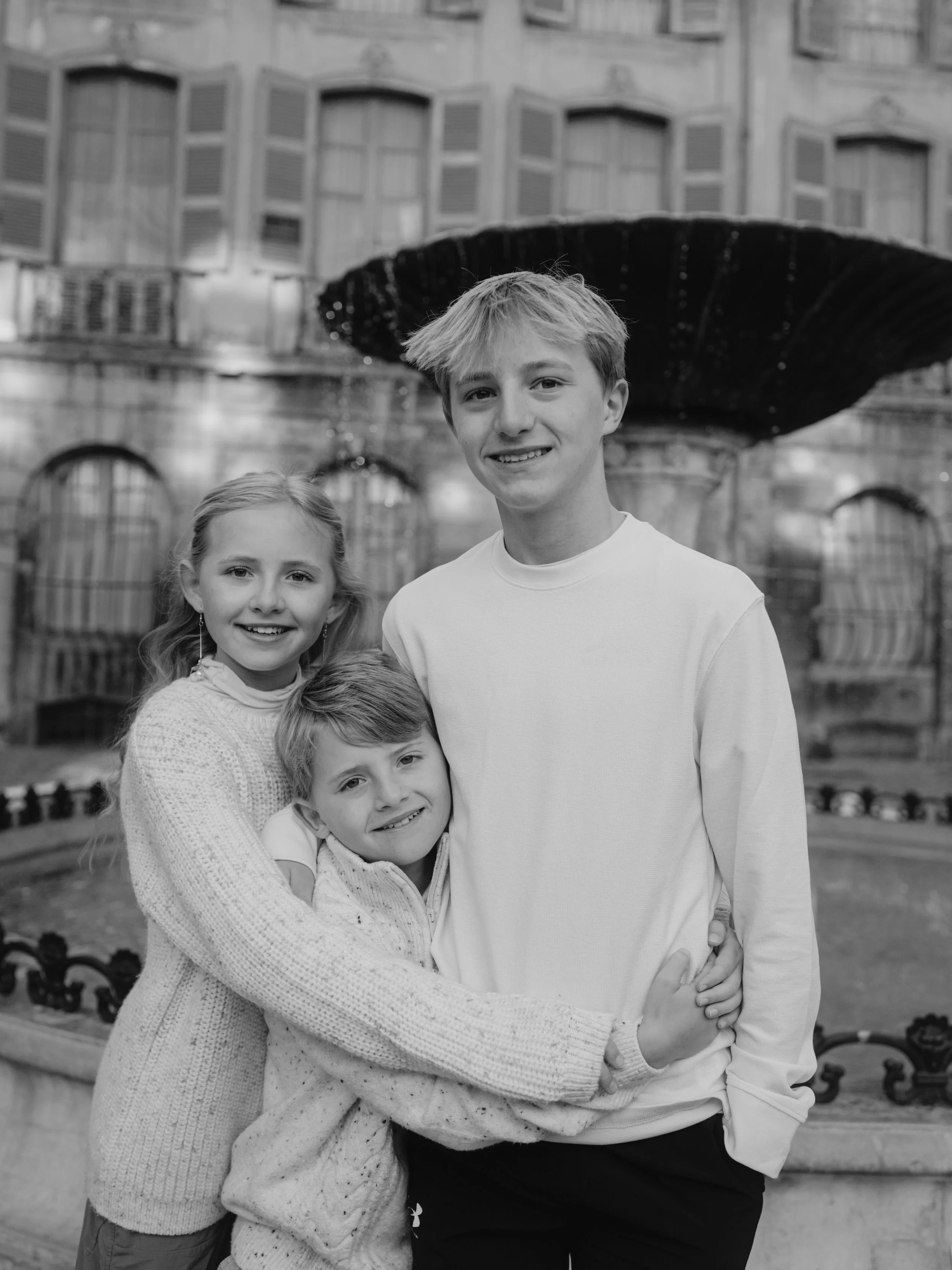 A group of three children, two boys and one girl, smiling and embracing each other in front of a fountain with a building in the background.