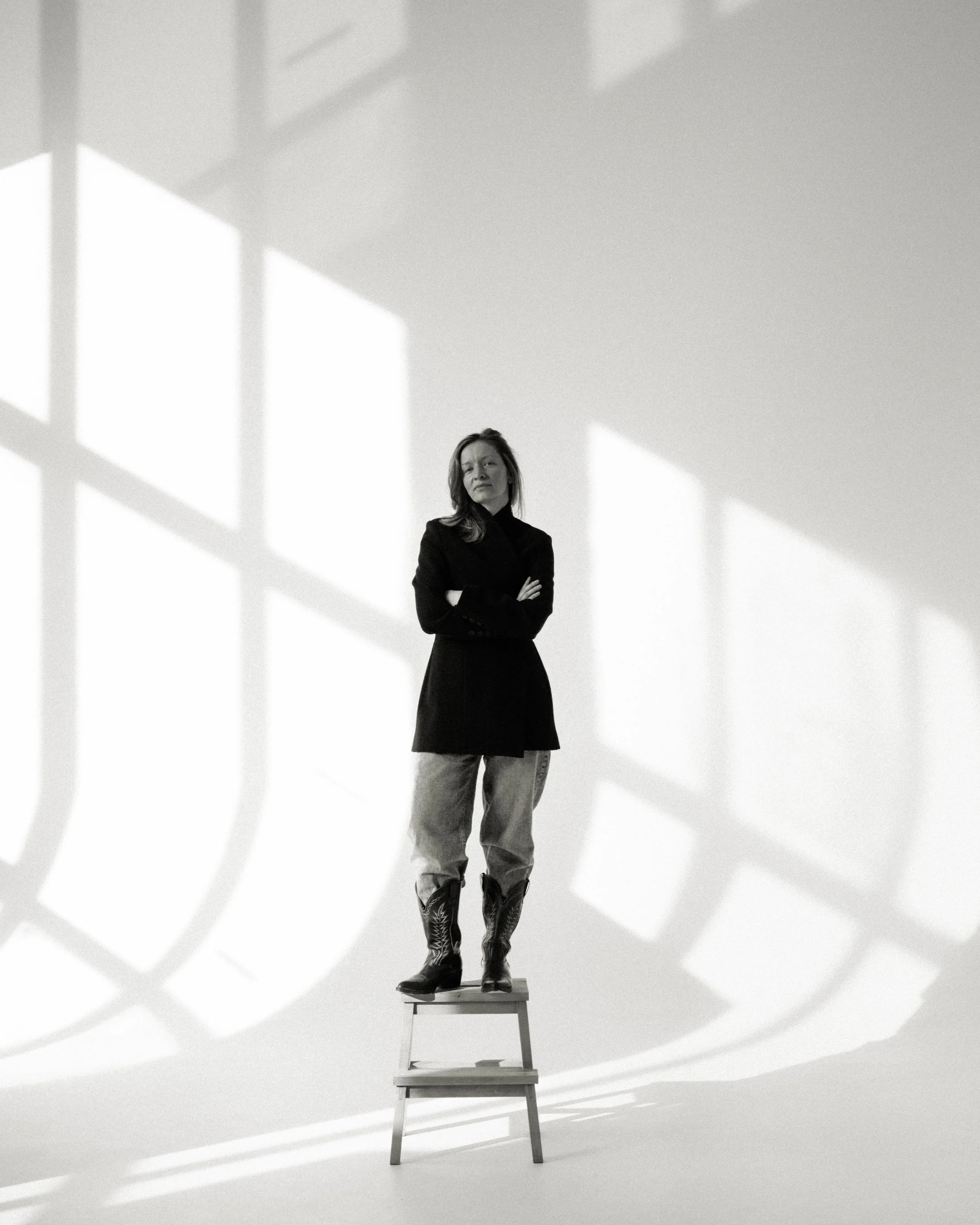 A woman standing on a step stool with crossed arms in front of a white wall with shadow patterns of window frames.