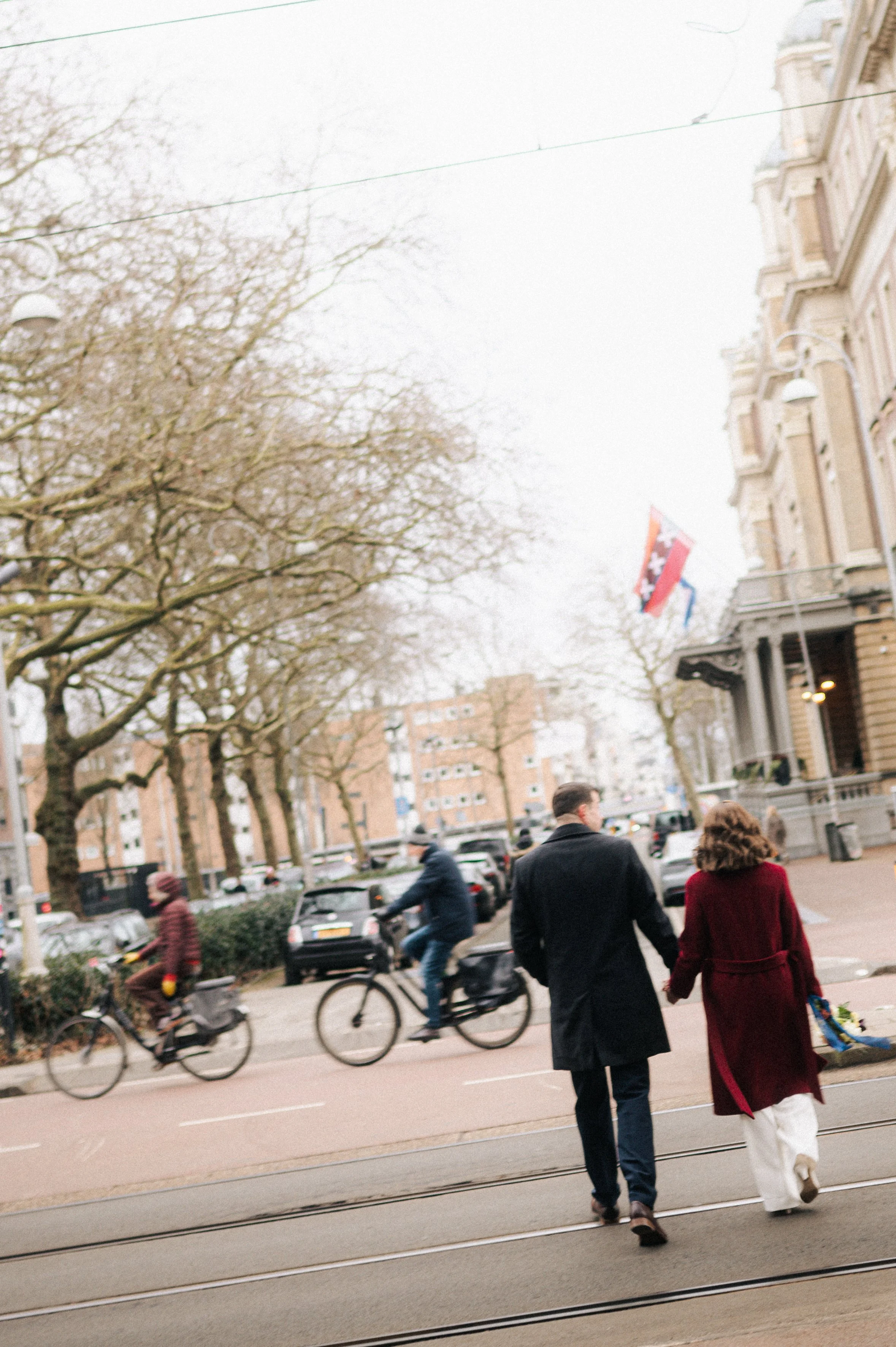 A couple walking hand in hand on a city street with people riding bicycles in the background, trees, and historic buildings.