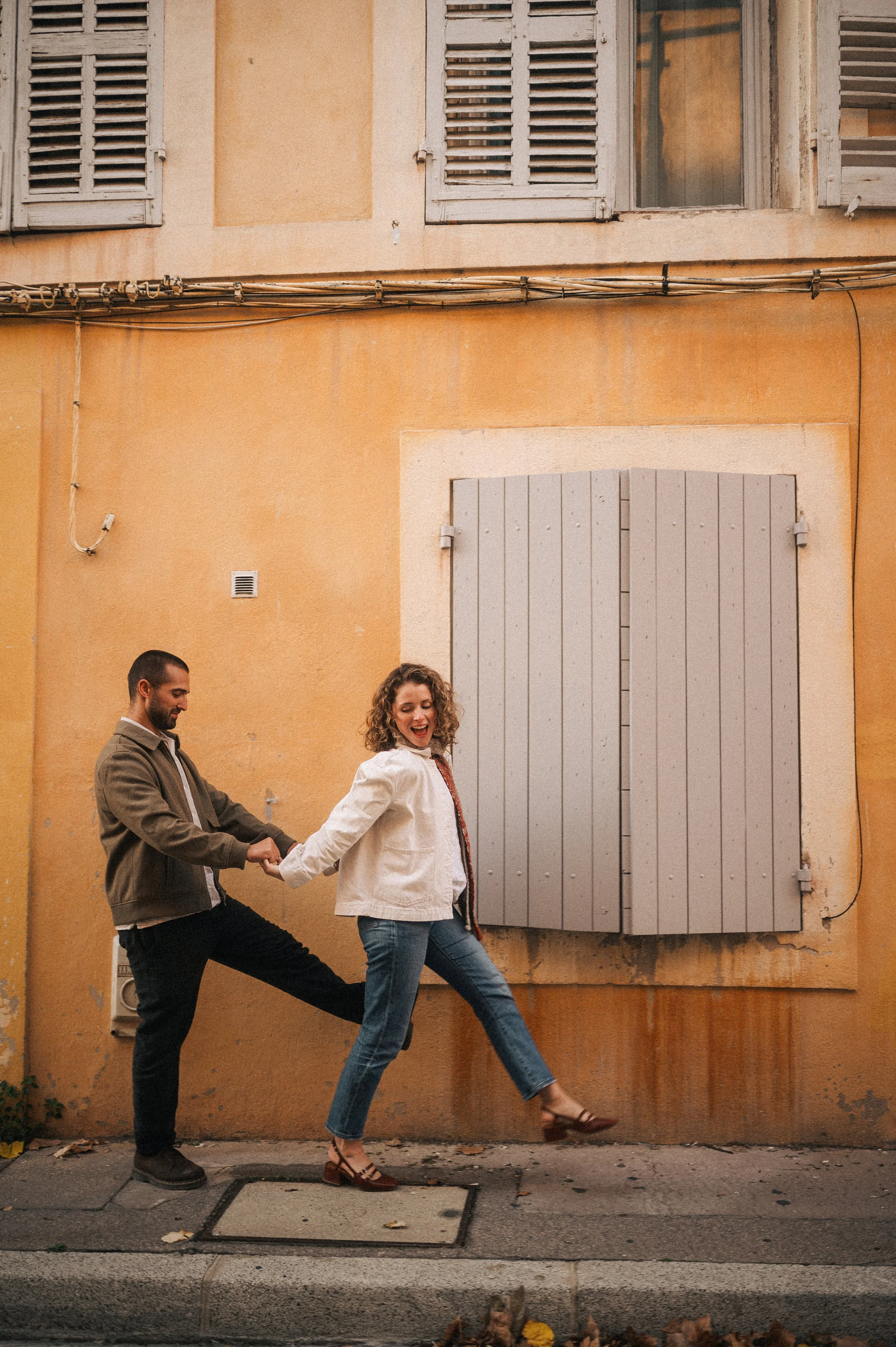 A man and woman holding hands and posing playfully on a city sidewalk, with the woman pretending to walk a step ahead while the man holds her hand, in front of an orange wall with closed gray shutters.