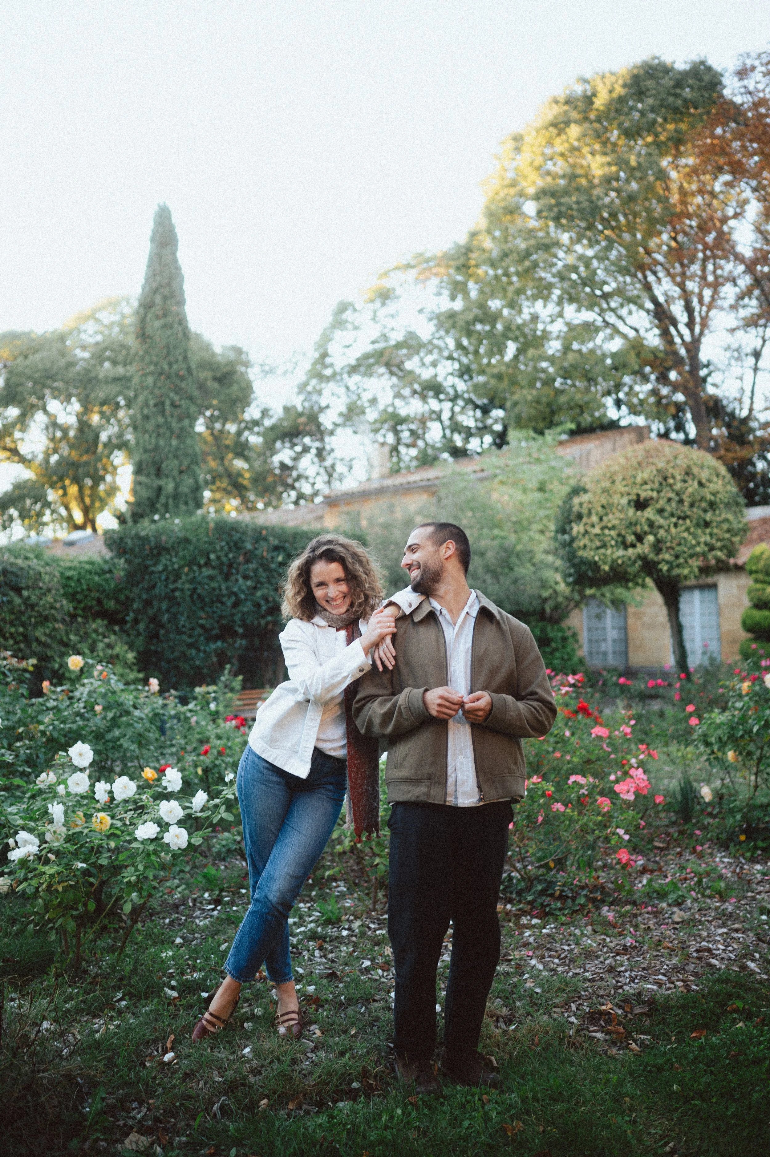 A smiling woman and man standing in a garden filled with colorful flowers, trees, and a house in the background, enjoying a moment together.