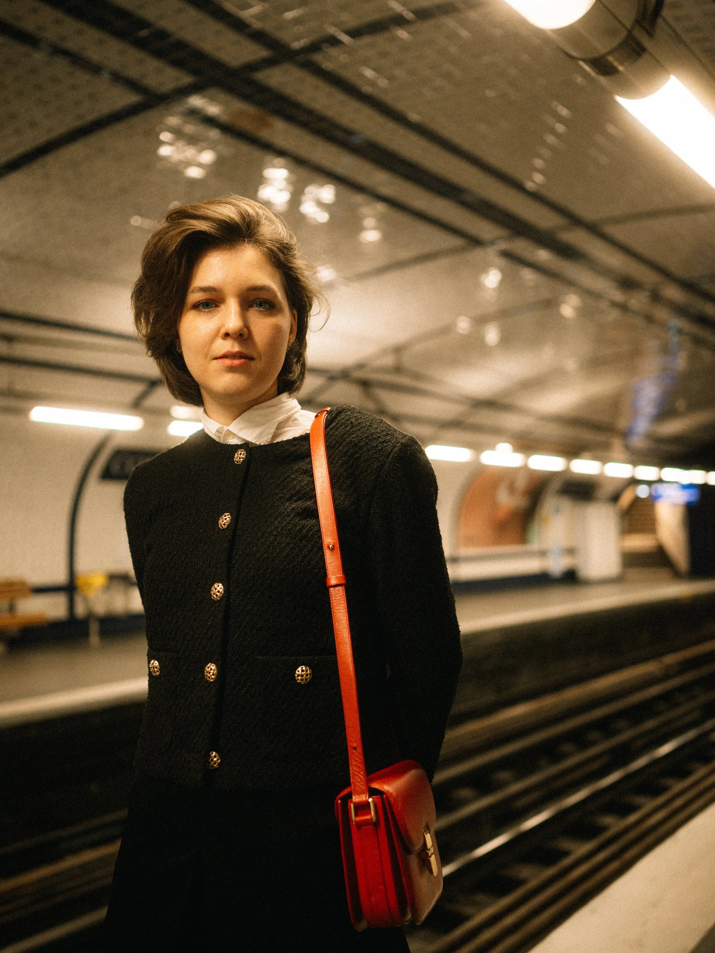 A woman with short brown hair wearing a black textured jacket with gold buttons, white shirt, and red handbag standing on a subway platform.