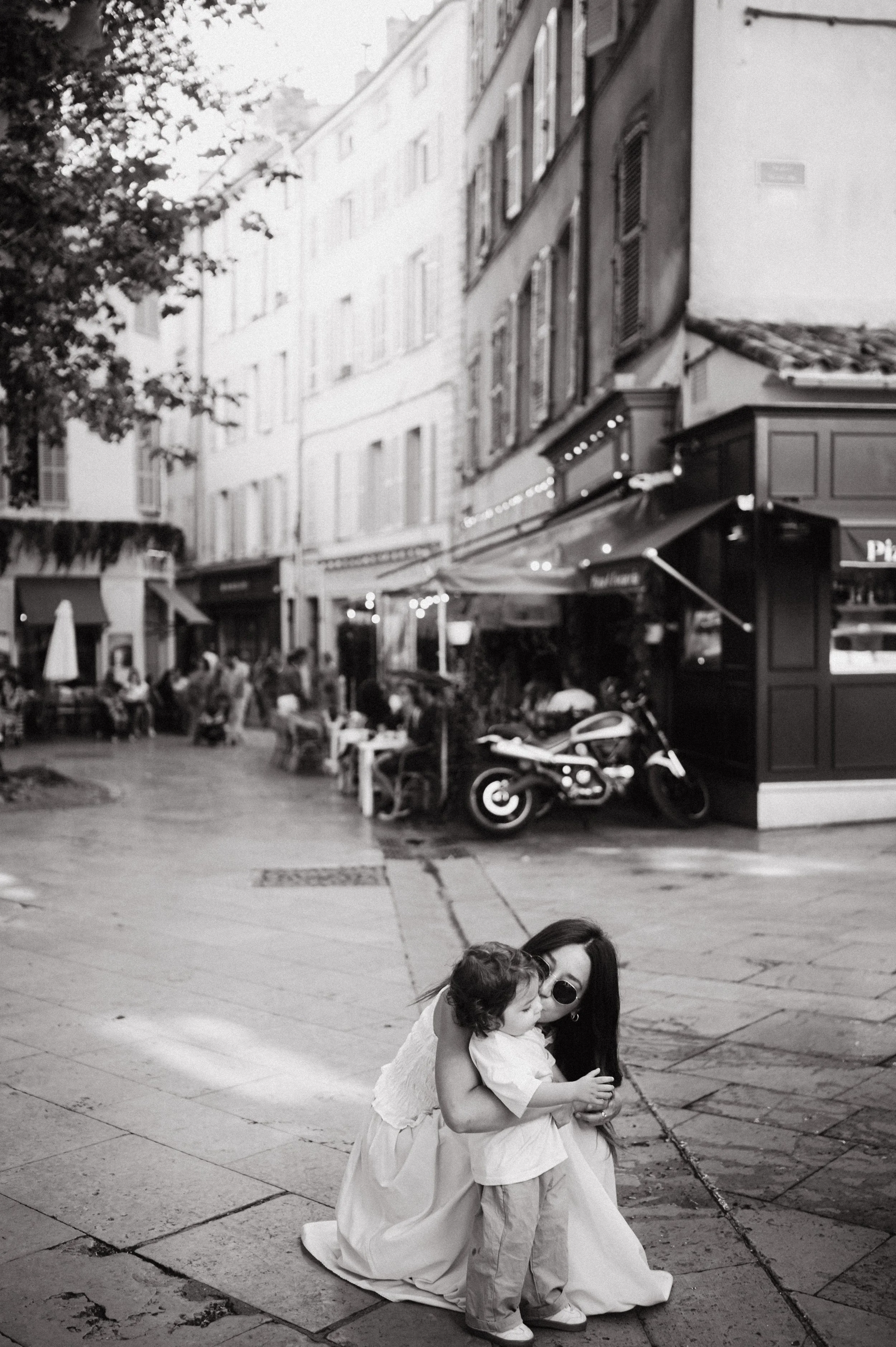 Woman wearing sunglasses and a long dress crouches on a city sidewalk, hugging a young child with dark hair and light-colored clothing, in an outdoor urban setting with buildings, tables, chairs, and a motorcycle in the background.