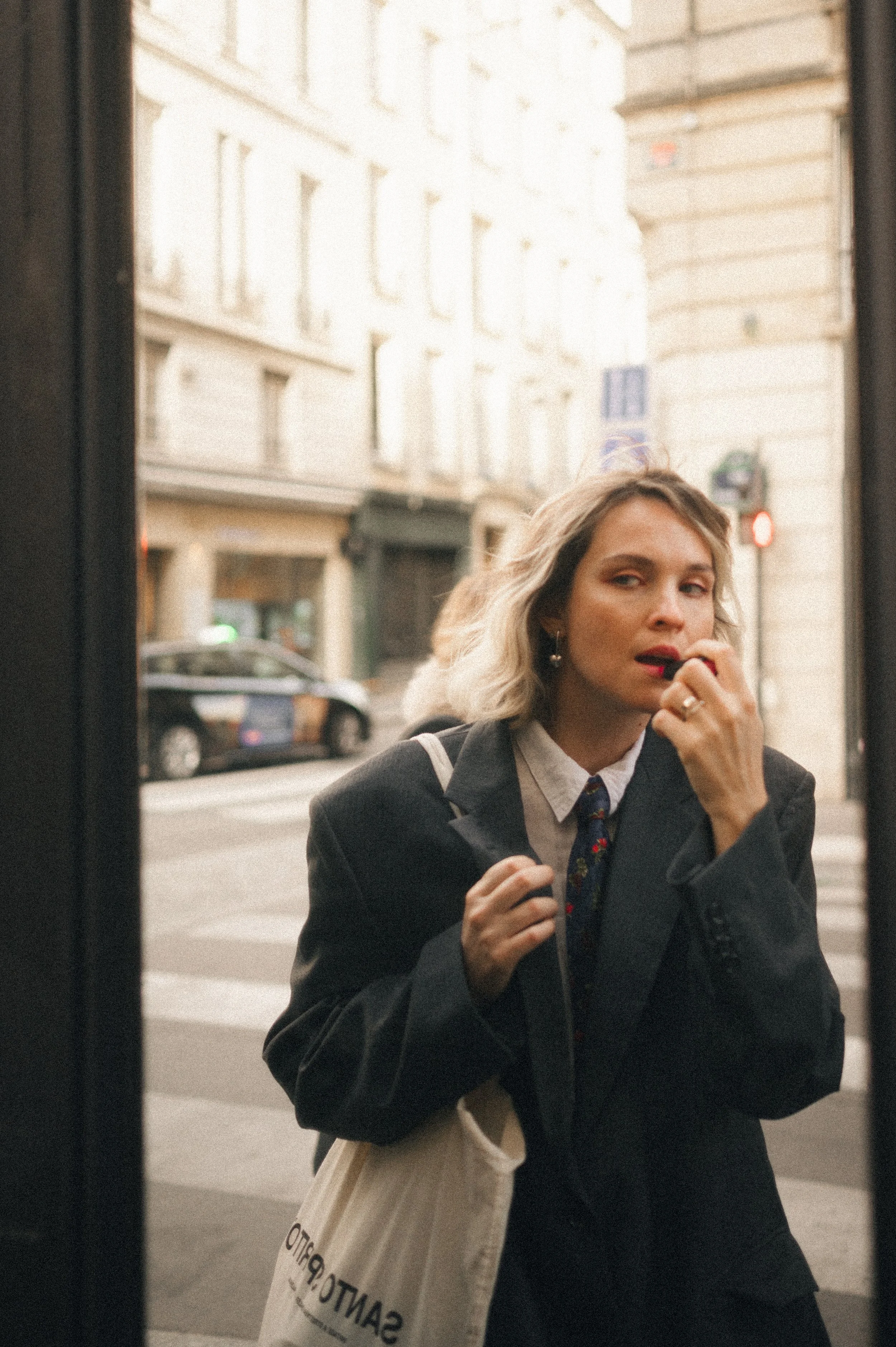 A woman with blonde hair, dressed in a black blazer and white shirt with a tie, looks into a mirror on a city street, holding a tote bag.