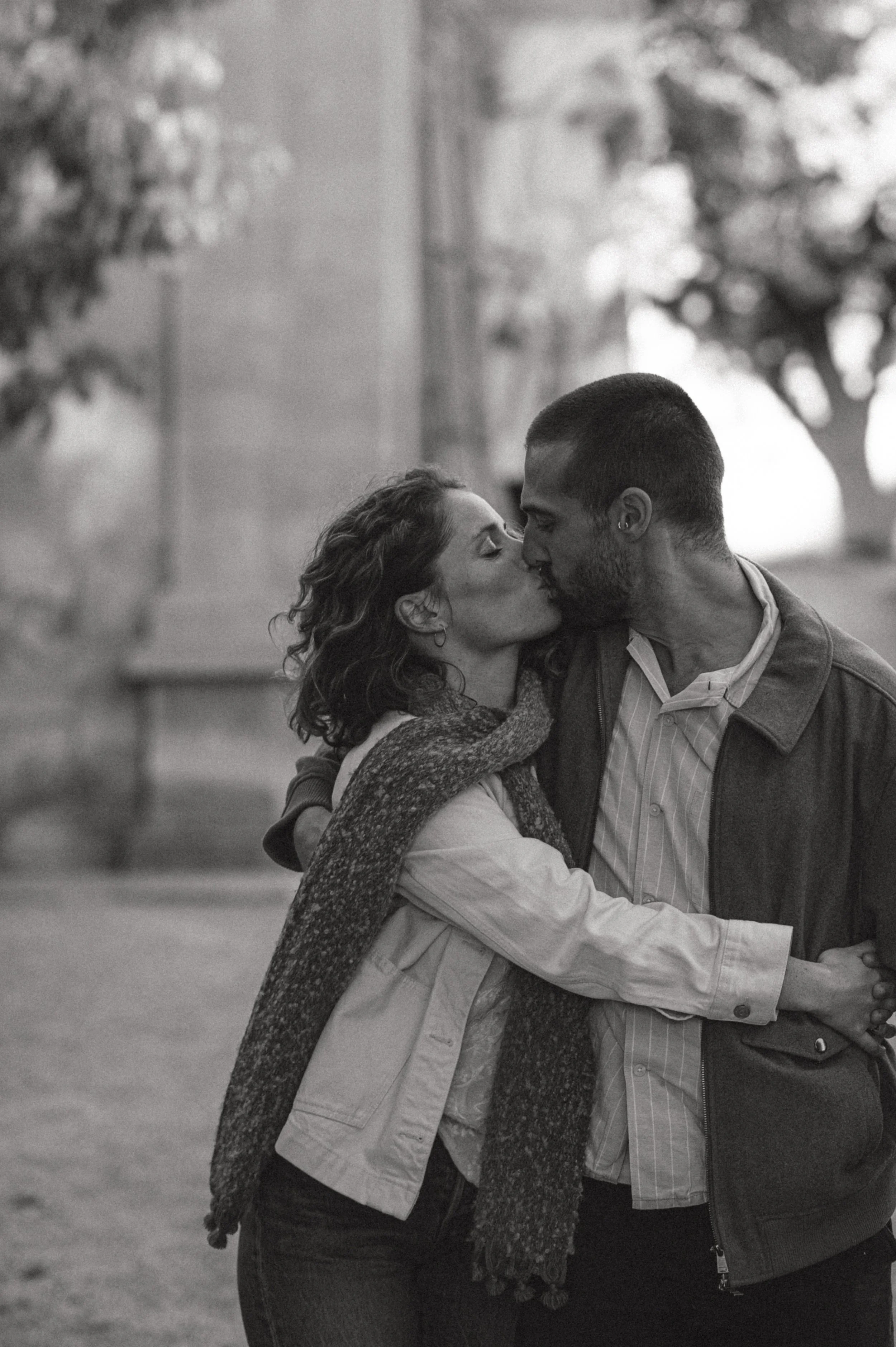 A black and white photo of a couple sharing a kiss outdoors, with trees and a bench in the background.