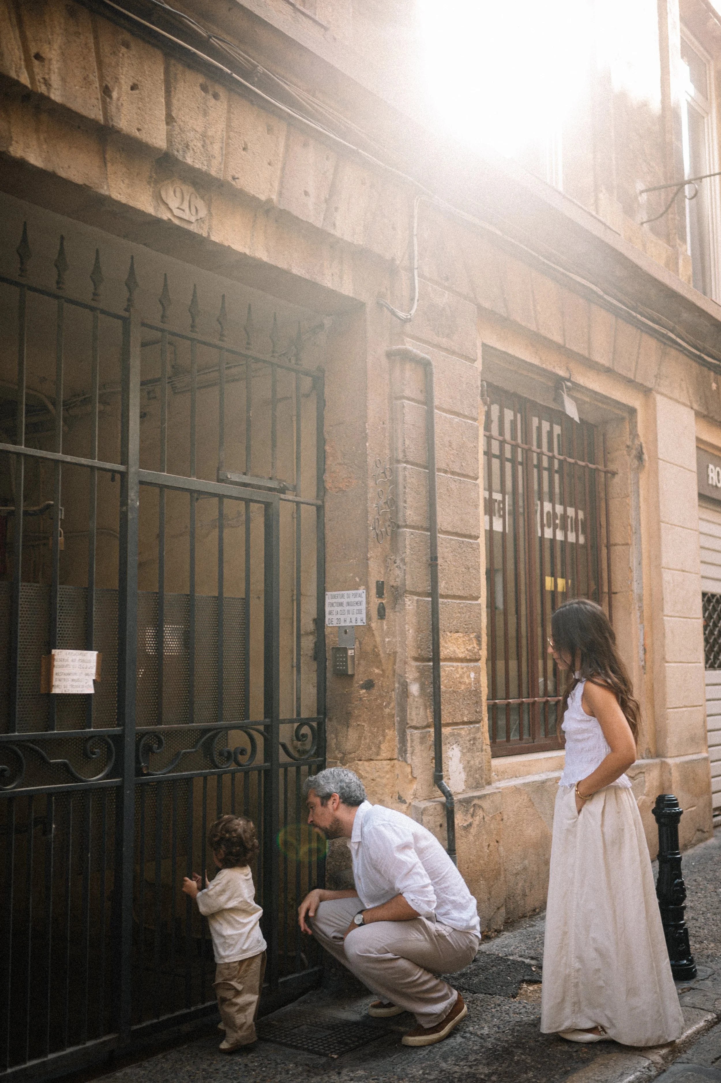 A man, woman, and young child with curly hair on a city street. The man is crouched near a gate, the child is standing with the man, and the woman is standing nearby. The building has stone walls with bars on the windows and a metal gate.