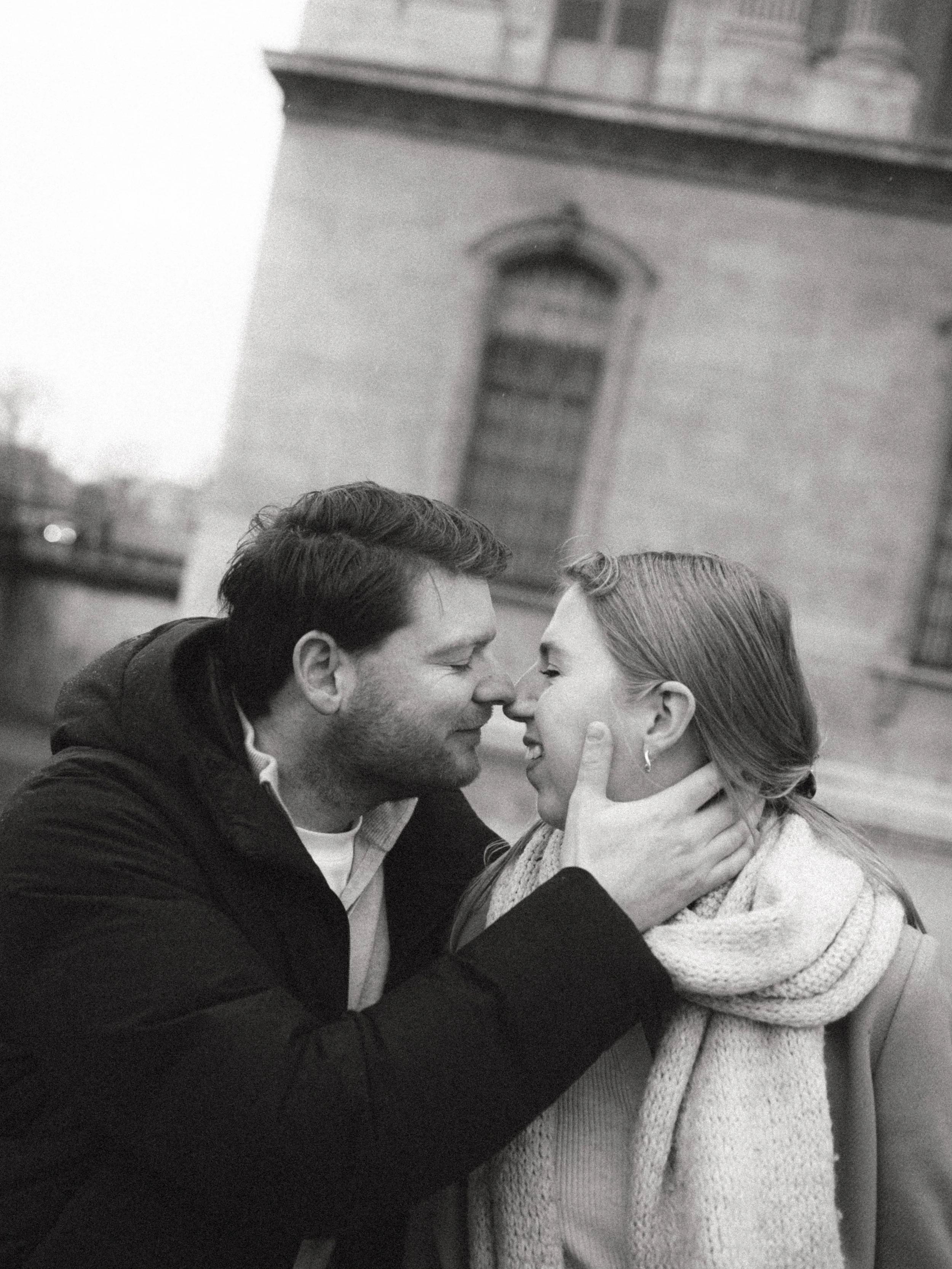 A black and white photo of a couple with closed eyes touching noses, outdoors, with a large brick building in the background.