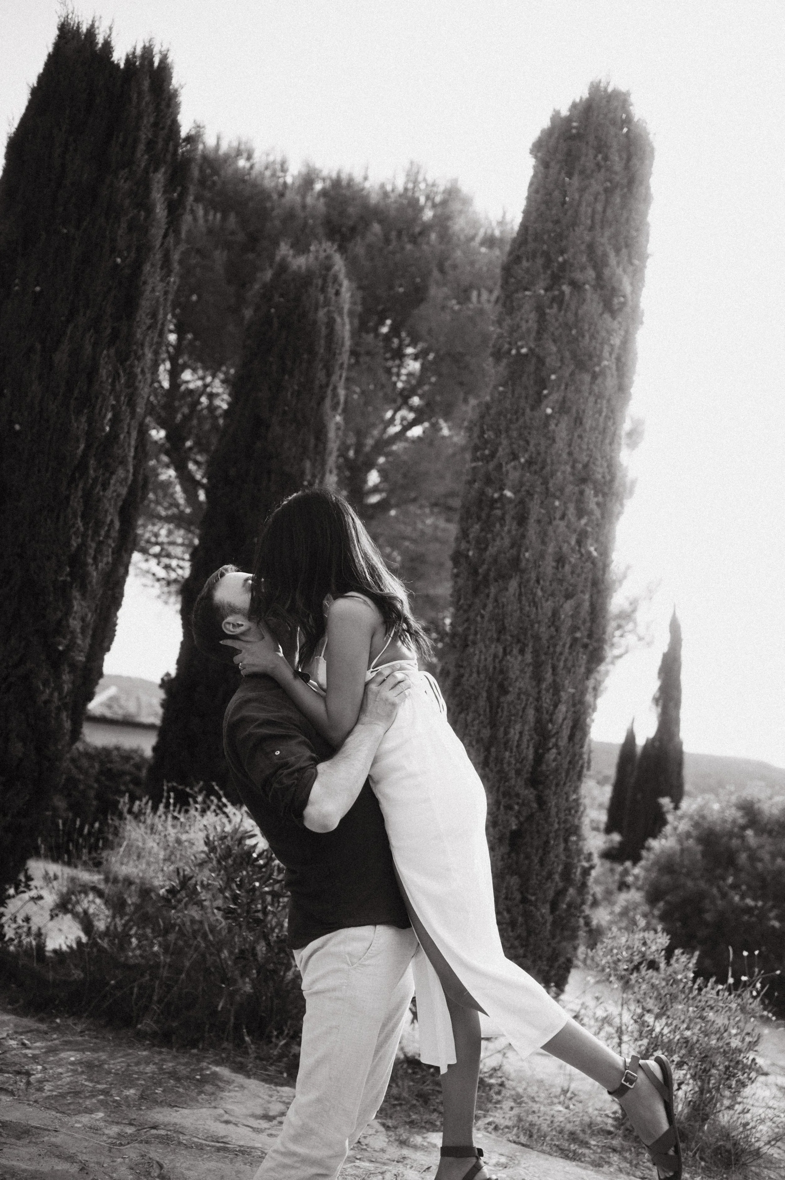 A couple in love, with the man lifting the woman, kissing outdoors near tall trees, black and white photo.