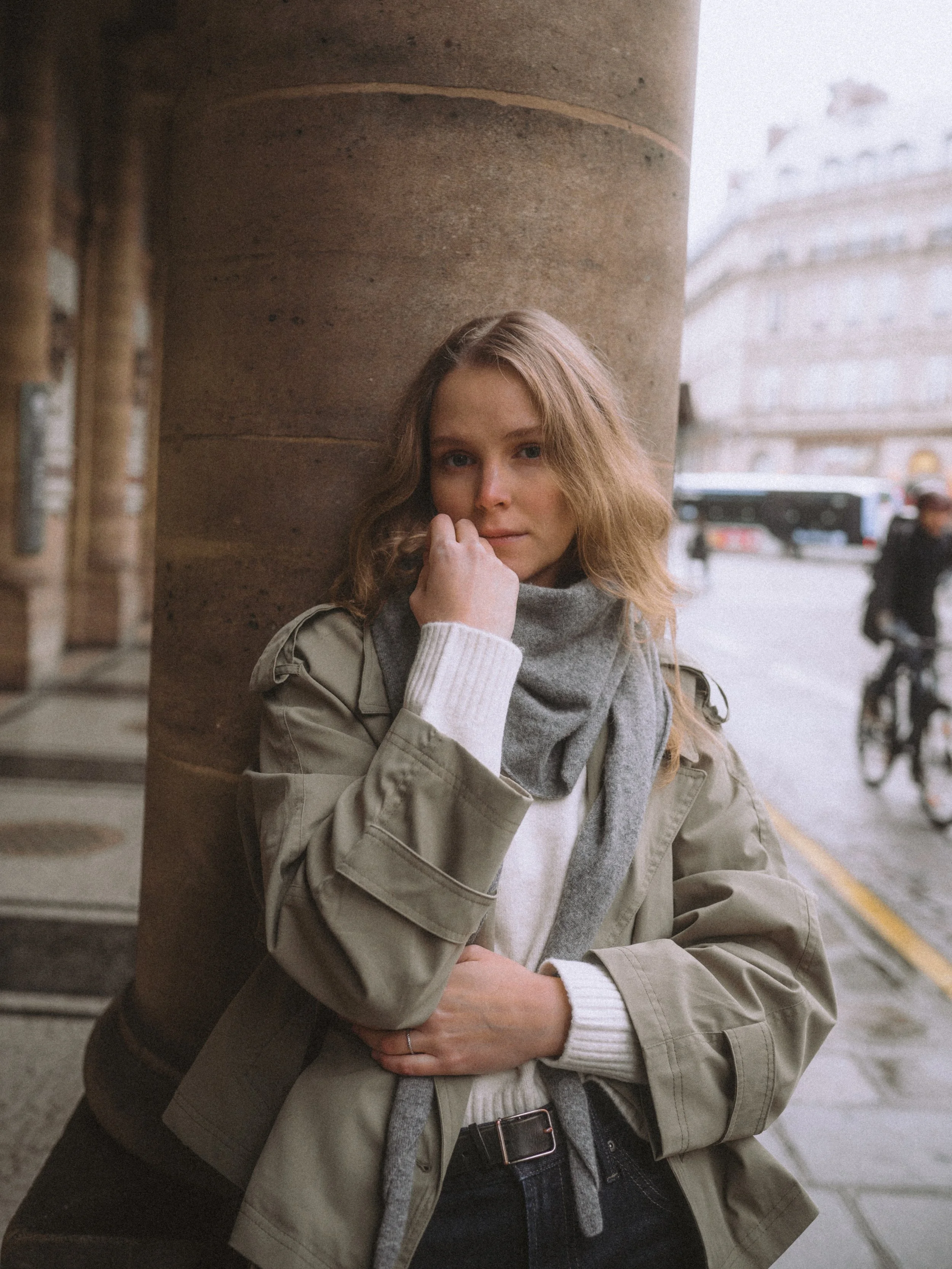 Young woman with curly hair leaning against a large stone column on a city street, wearing a beige jacket, white sweater, gray scarf, and black jeans.