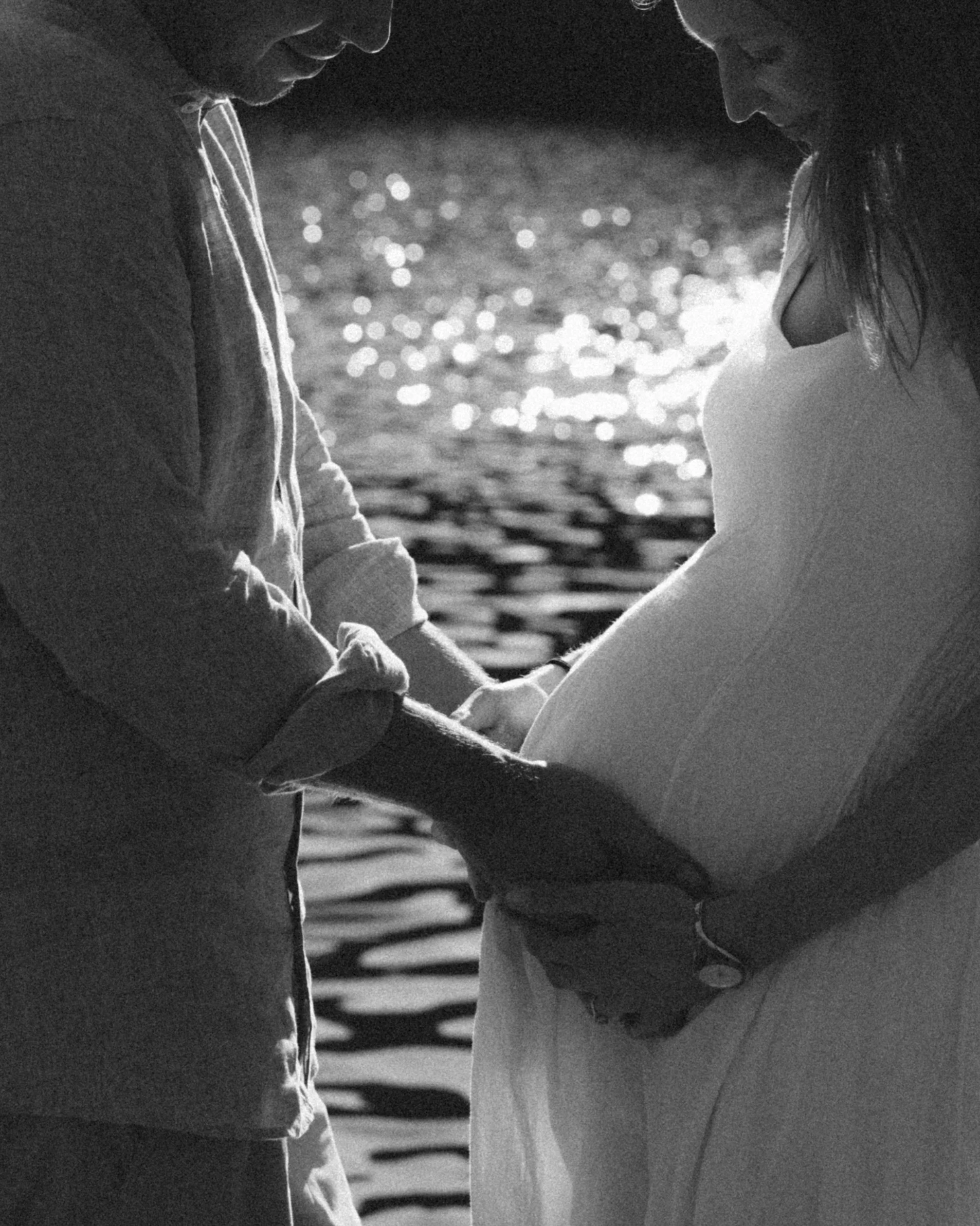 A pregnant woman in a white dress holds her belly as a partner gently touches her stomach, standing near a body of water with sunlight reflecting off the surface.