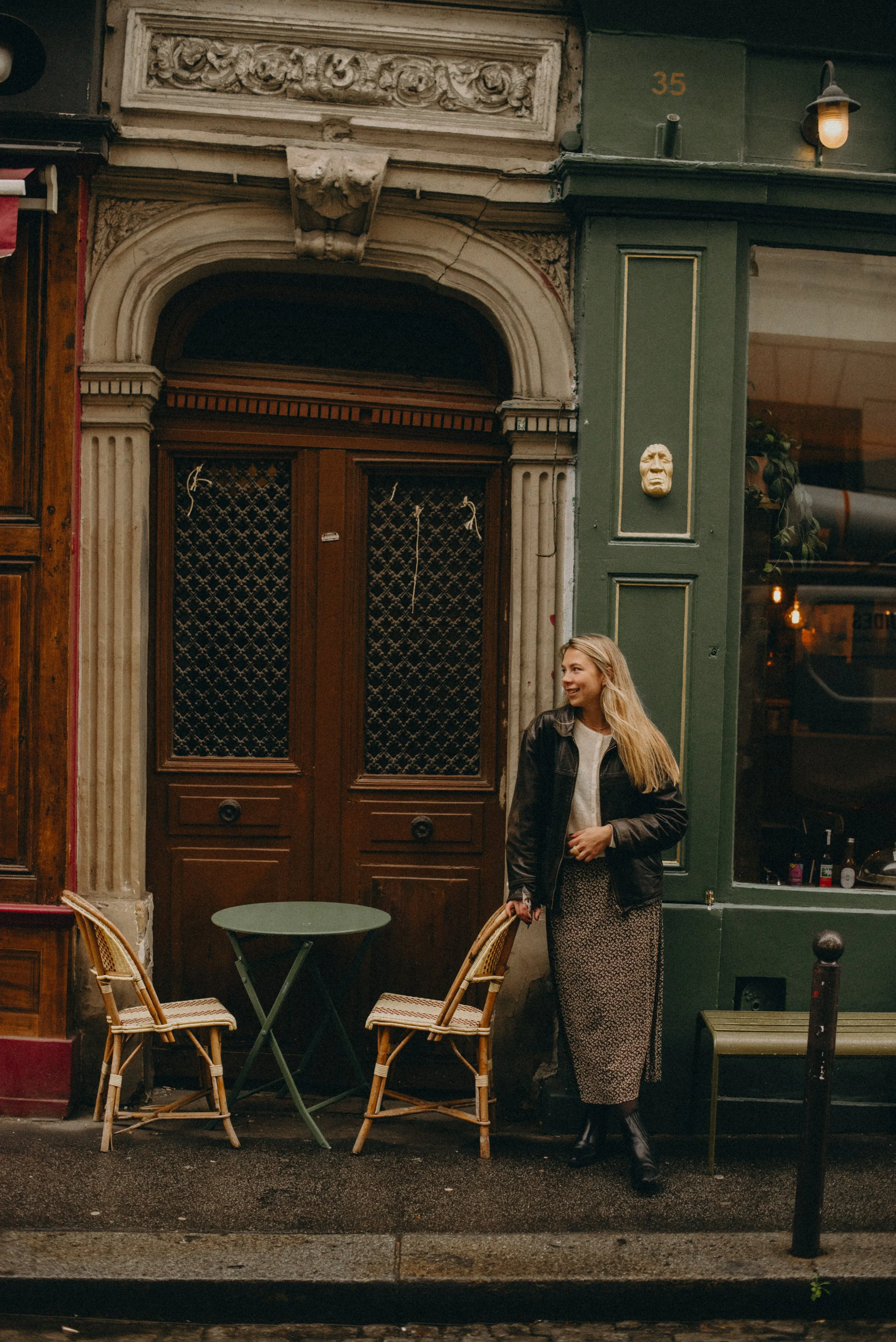 A woman with long blonde hair smiling and standing outside a cafe, holding a smartphone in one hand. She is wearing a black leather jacket, a gray top, a patterned skirt, and black boots. There is a small round green table with two chairs beside her. The cafe has a green and wooden exterior with decorative details around the door.