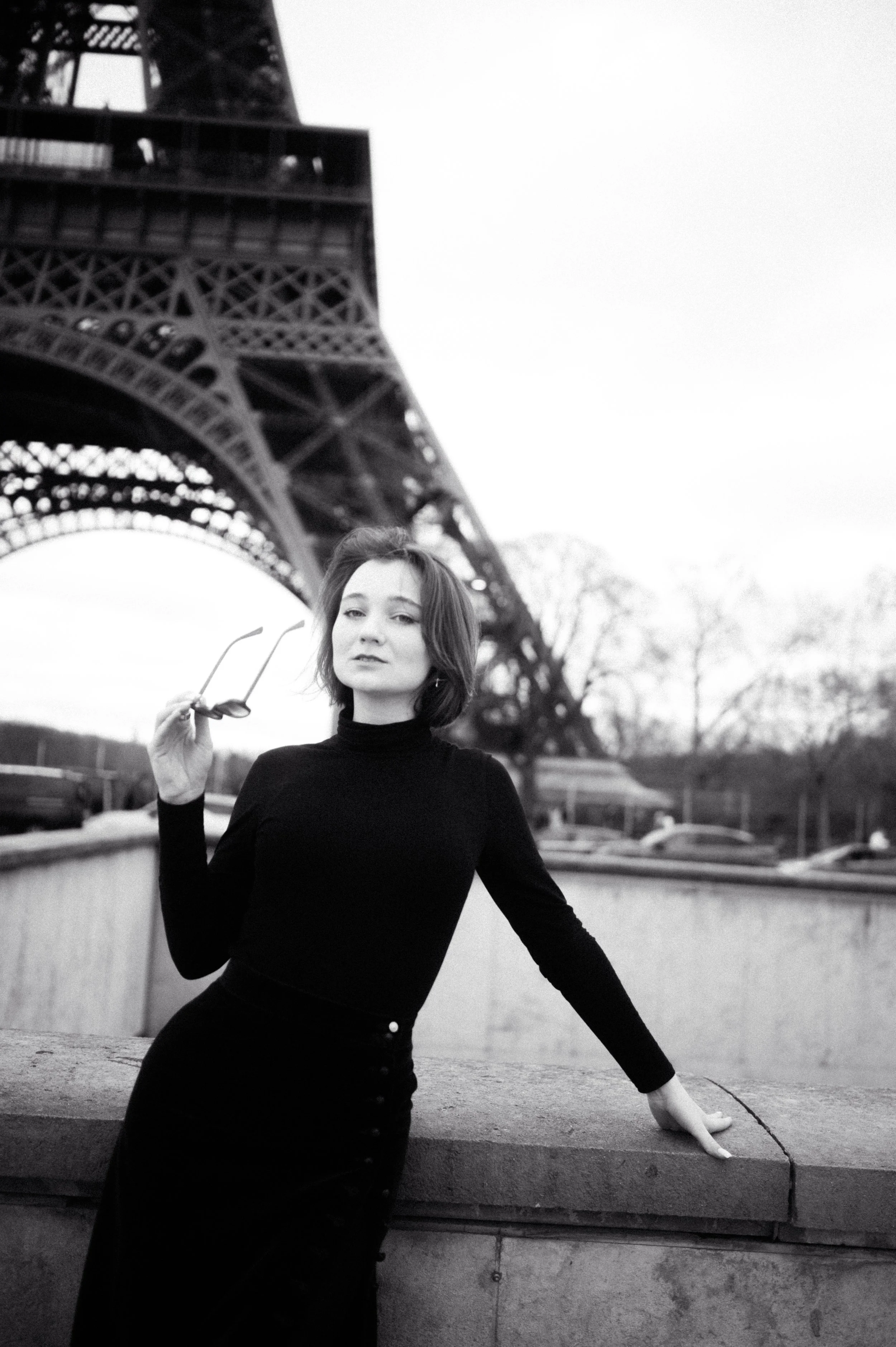 A woman in a black long-sleeve top holding sunglasses, leaning on a ledge near the Eiffel Tower in Paris, France.