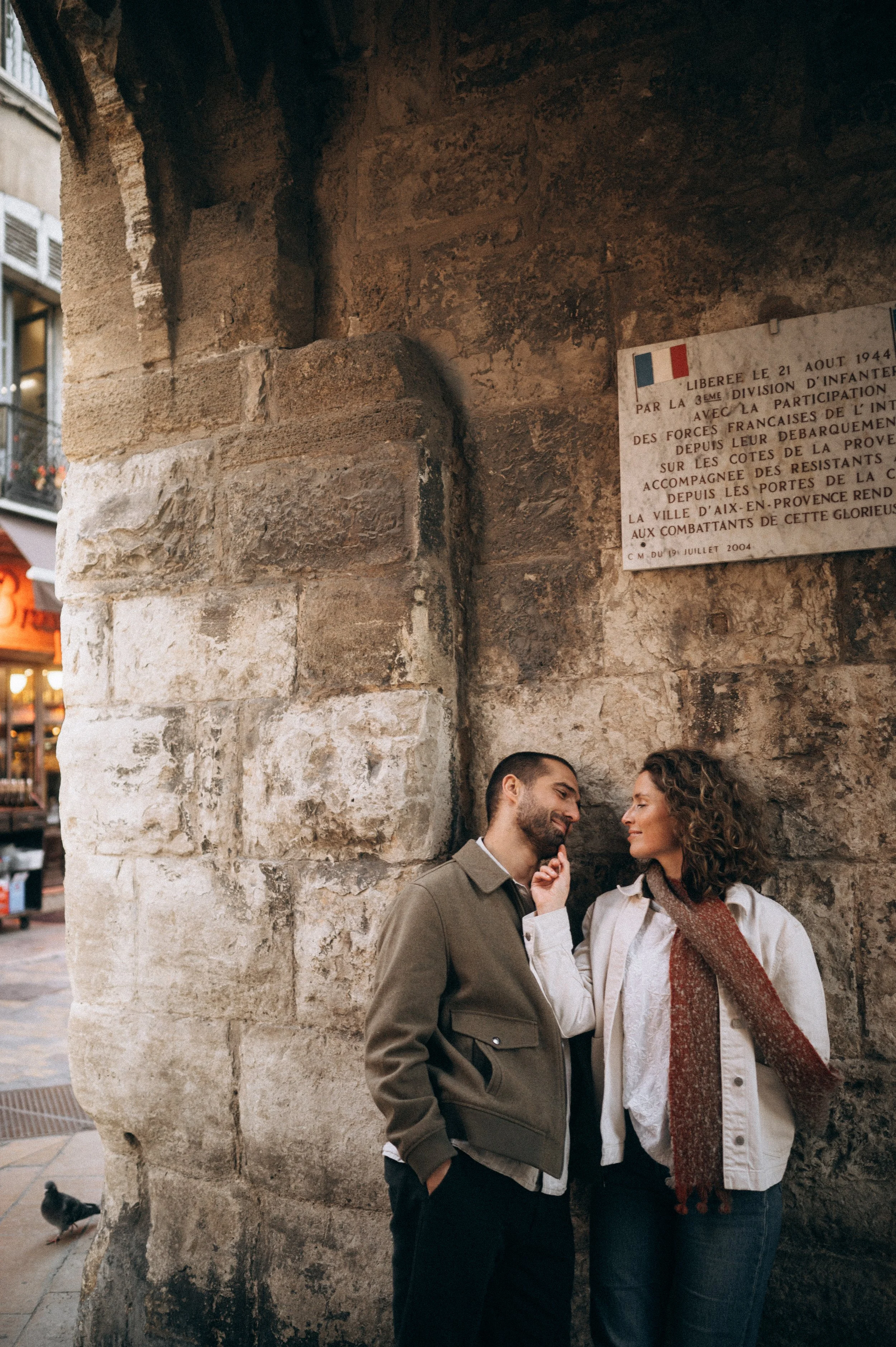 A couple leaning against a stone wall in a historic European city, sharing a tender moment, with a plaque and buildings visible in the background.