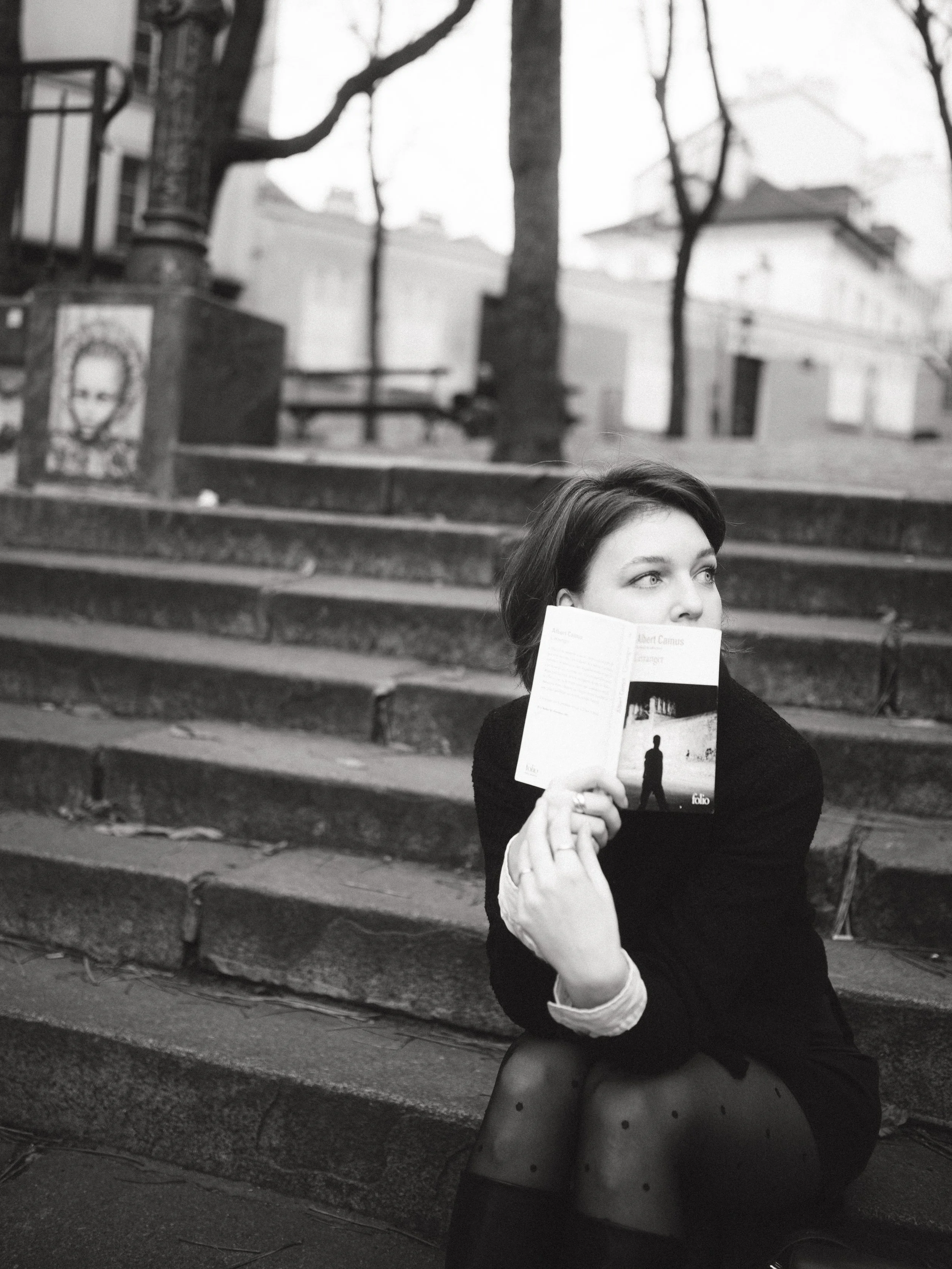 A woman sitting on outdoor steps holding a book in front of her face, with her head turned to the side, in a city park setting with trees and buildings in the background, black and white photo.