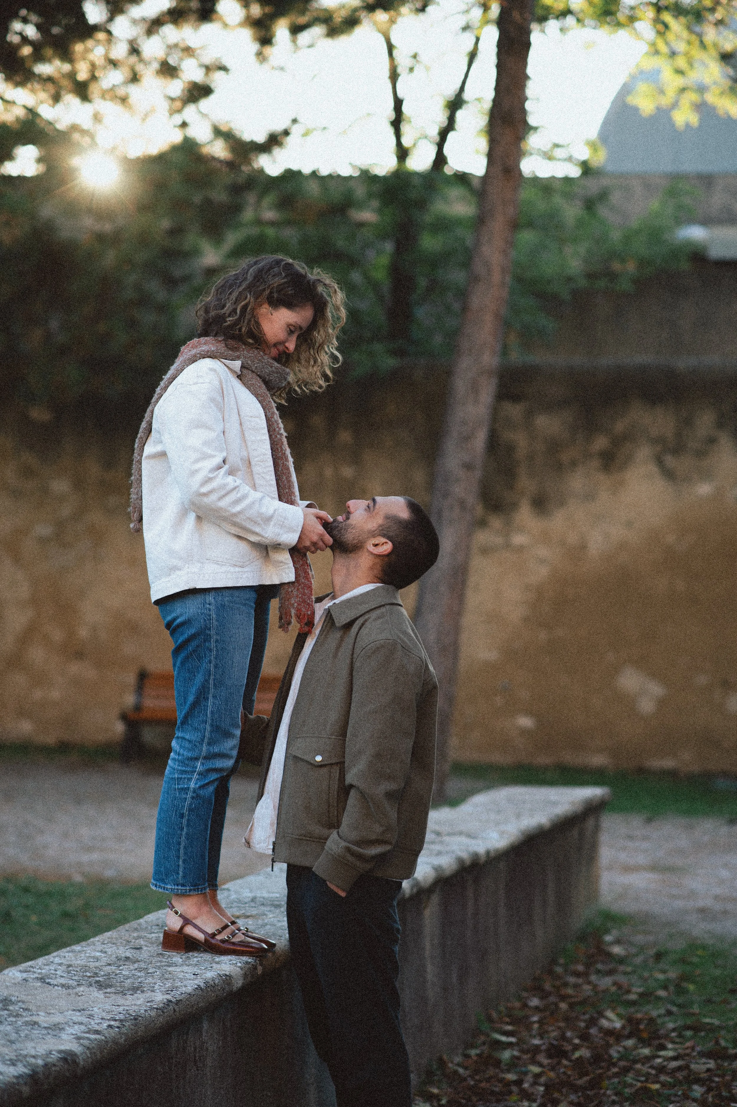 A woman standing on a stone ledge, leaning slightly forward, is gently touching the chin of a man who is standing in front of her, looking up at her. They are outdoors during sunset with trees and a building in the background.