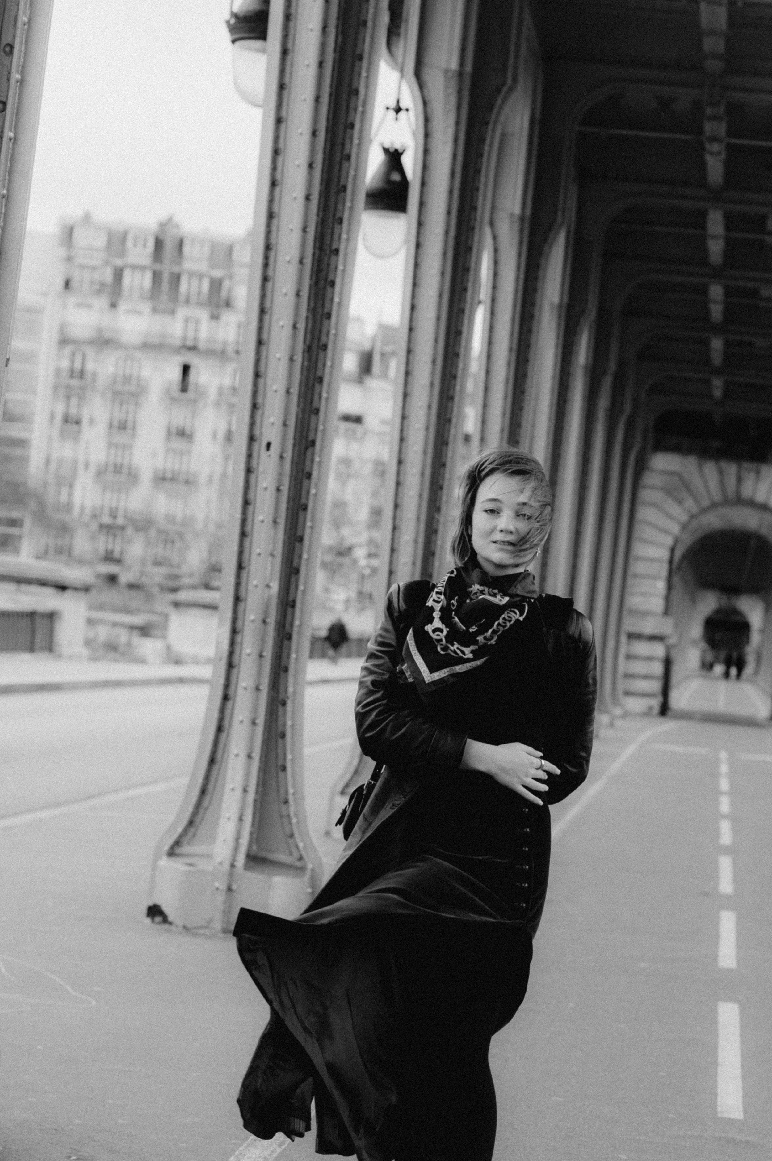 A woman standing on a bridge with city buildings in the background, wind blowing her hair, wearing a leather jacket and a scarf, in black and white.