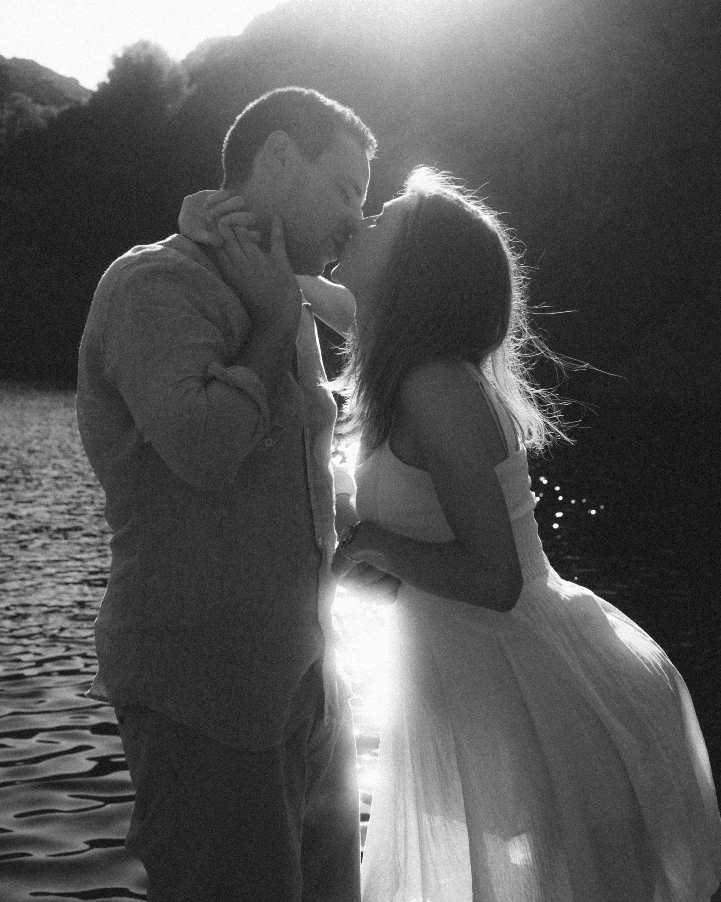 A black and white photo of a couple kissing near a body of water during sunset.