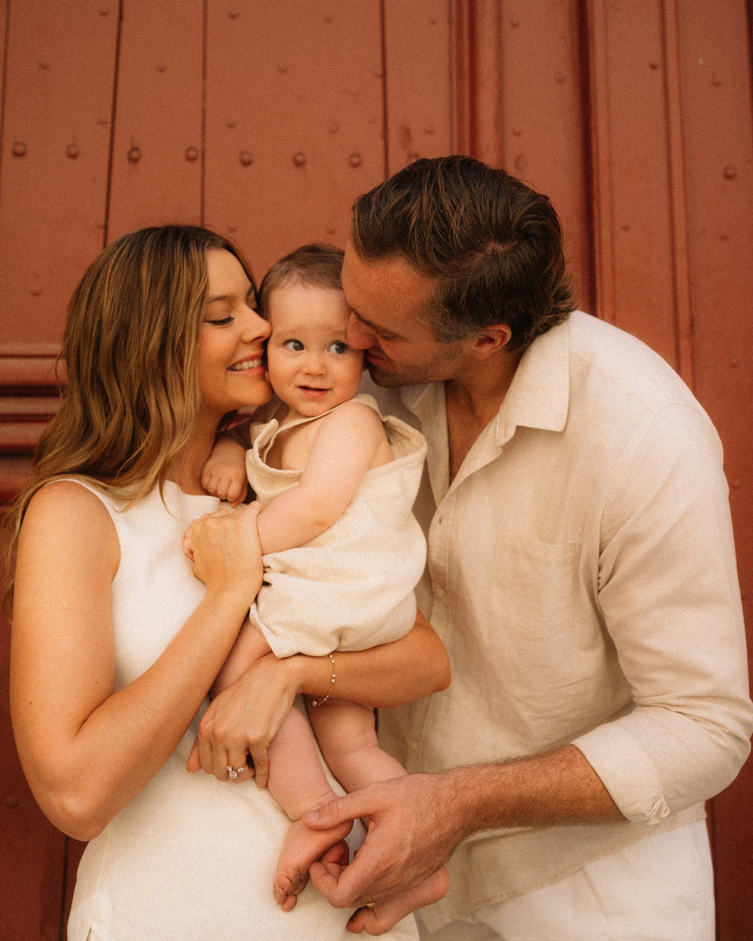 A family of three, with a woman, a man, and a baby, enjoying a close moment against a wooden wall background. The woman is holding the baby, and the man is leaning in to kiss the baby's cheek.