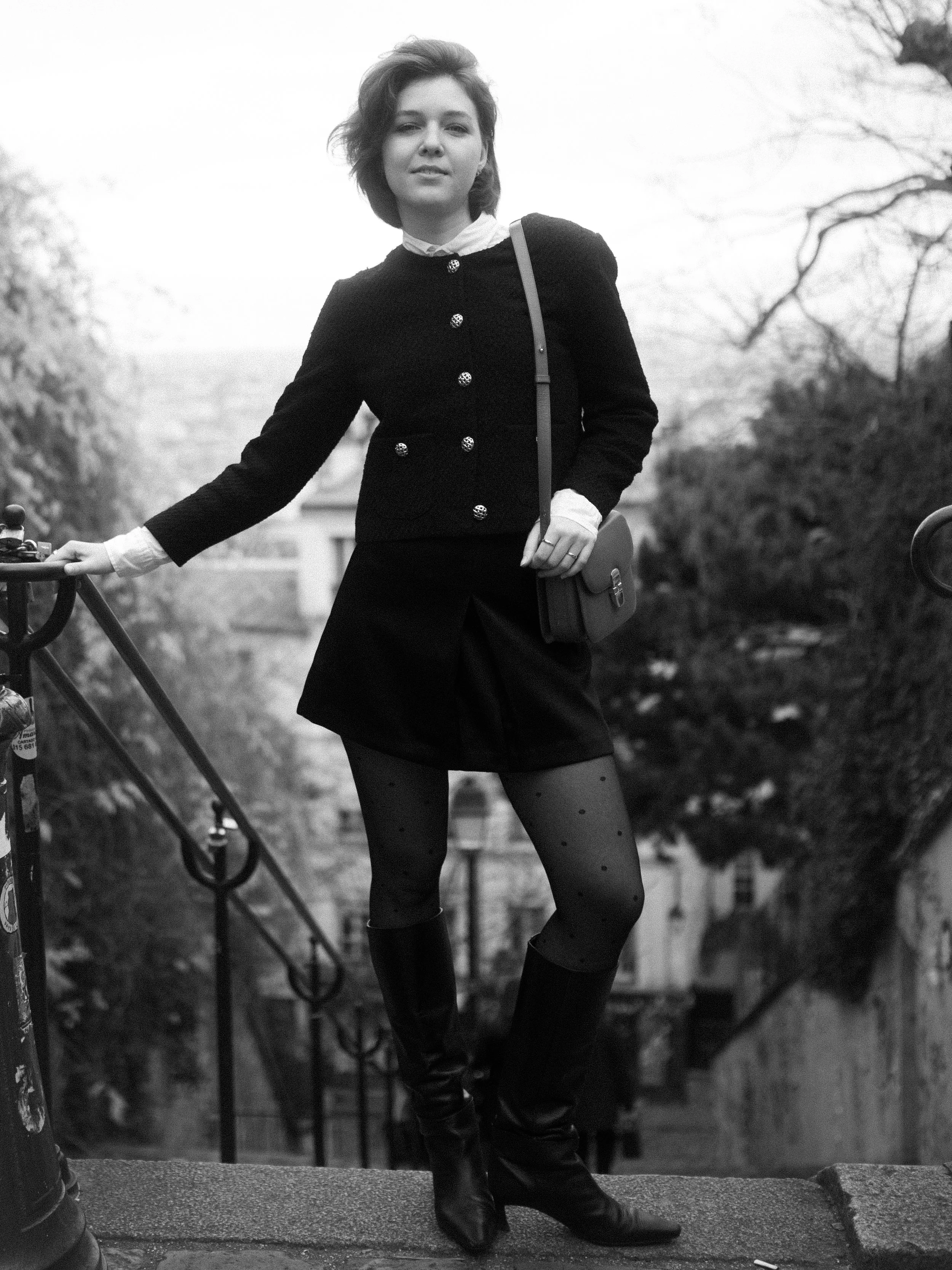 A woman standing outdoors on a staircase, wearing a dark jacket, skirt, patterned tights, and high boots, with trees and buildings in the background.